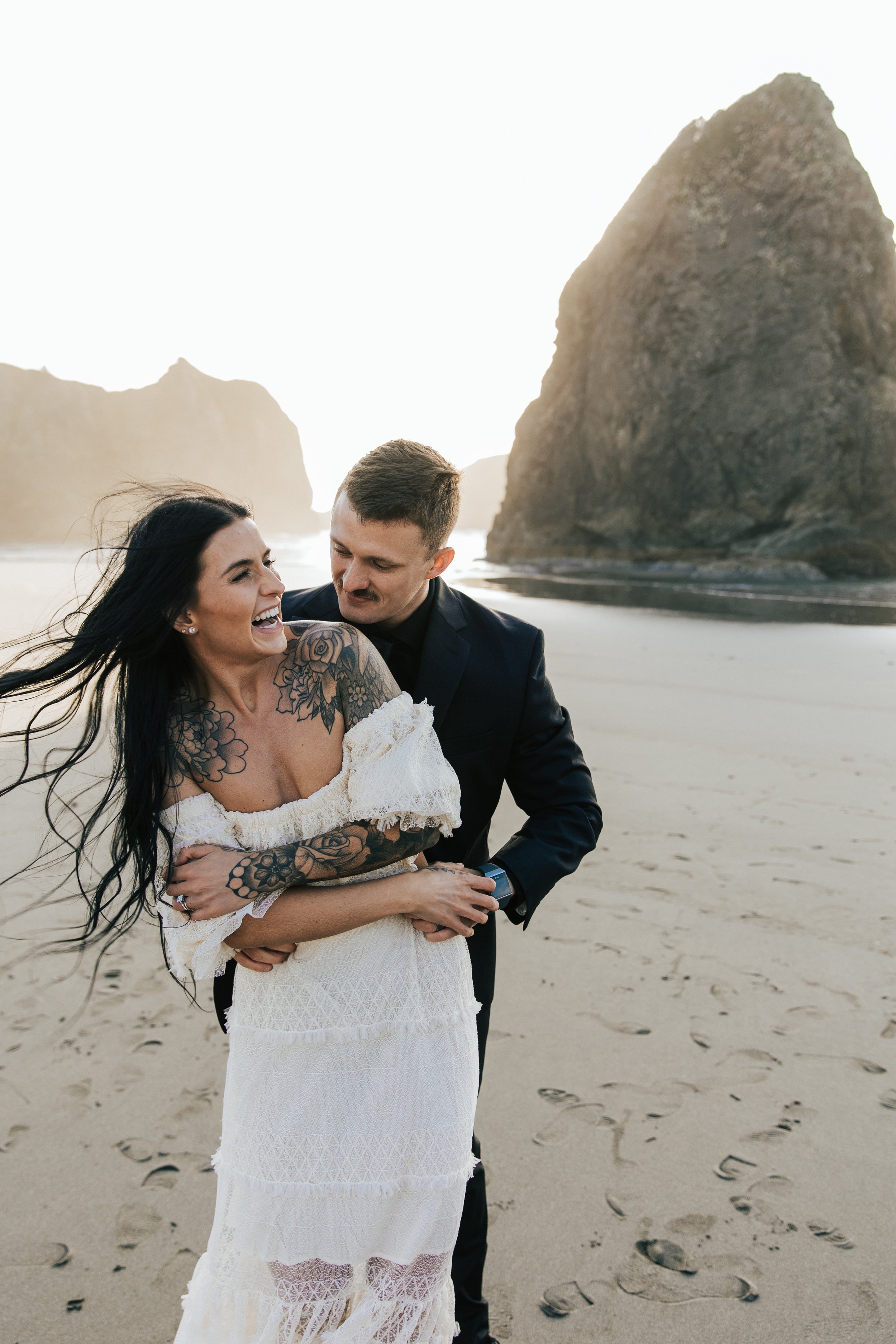  Oregon coast elopement. Couple elopes on the beach with the sun shining behind them. Sun rays behind rocks. Beach with rocks and haystacks. Southern Oregon. Golden hour photos after elopement ceremony. Couple laughs together on the beach. #oregoncoa