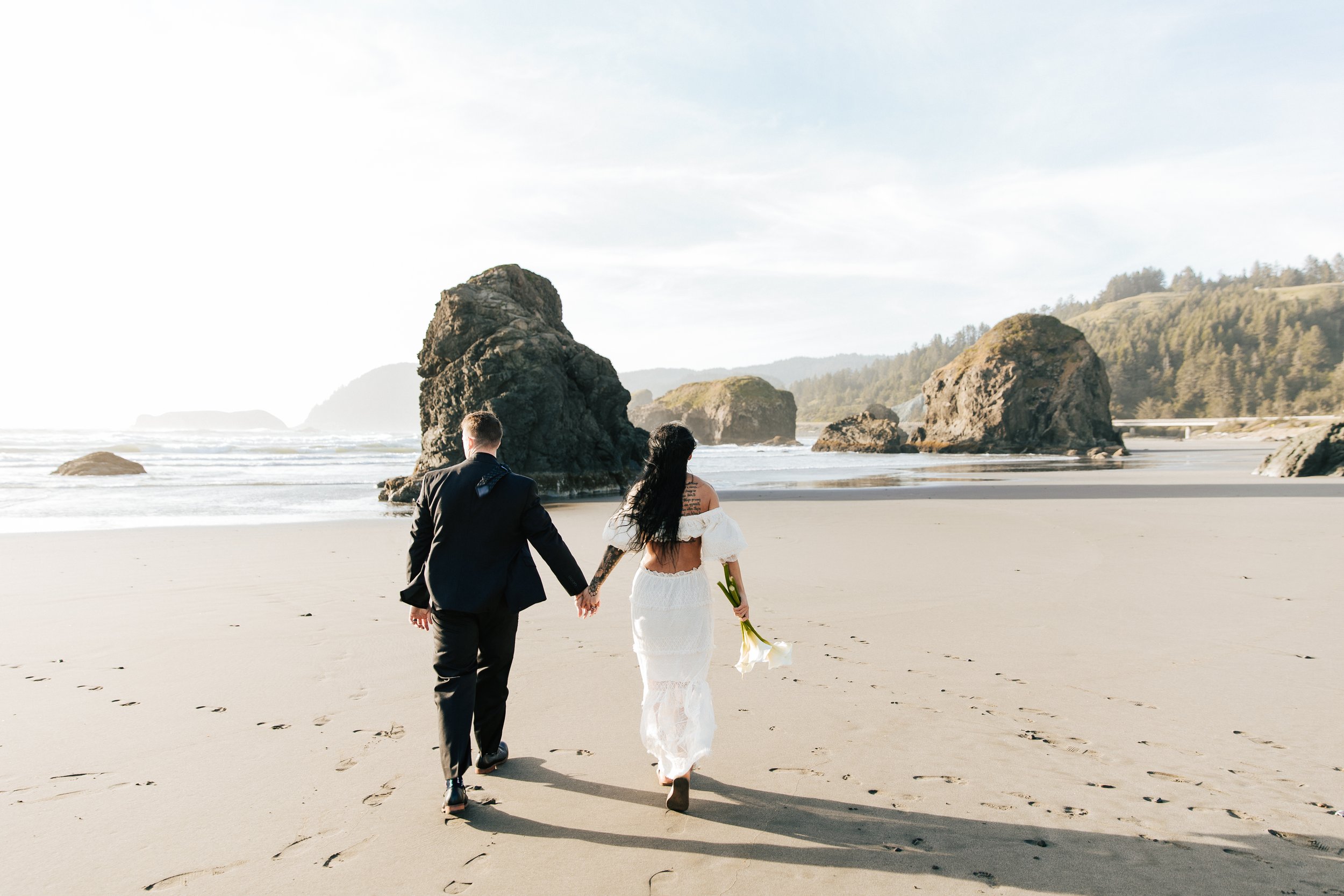  Oregon coast elopement. Couple elopes on the beach with the sun shining behind them. Sun rays behind rocks. Beach with rocks and haystacks. Southern Oregon. Brookings, Oregon. Samuel H Boardman elopement. Couple walks along the beach holding hands. 