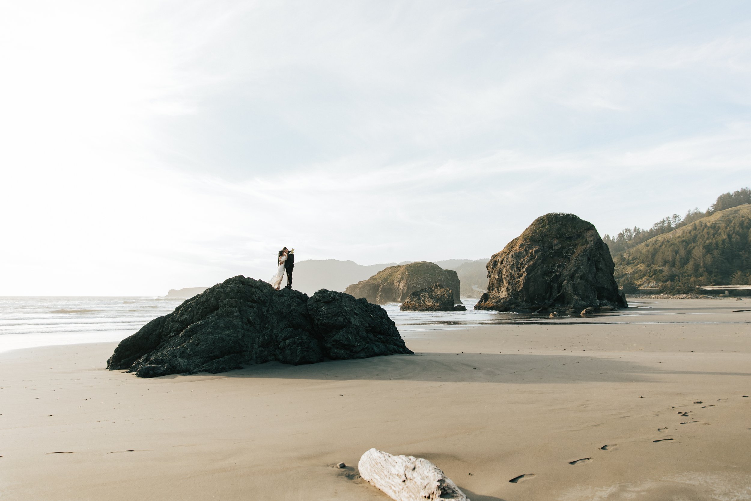  Oregon coast elopement. Couple elopes on the beach with the sun shining behind them. Sun rays behind rocks. Beach with rocks and haystacks. Southern Oregon. Couple stands up on a seastack or big rock far away on the beach. Brookings, Oregon. Samuel 
