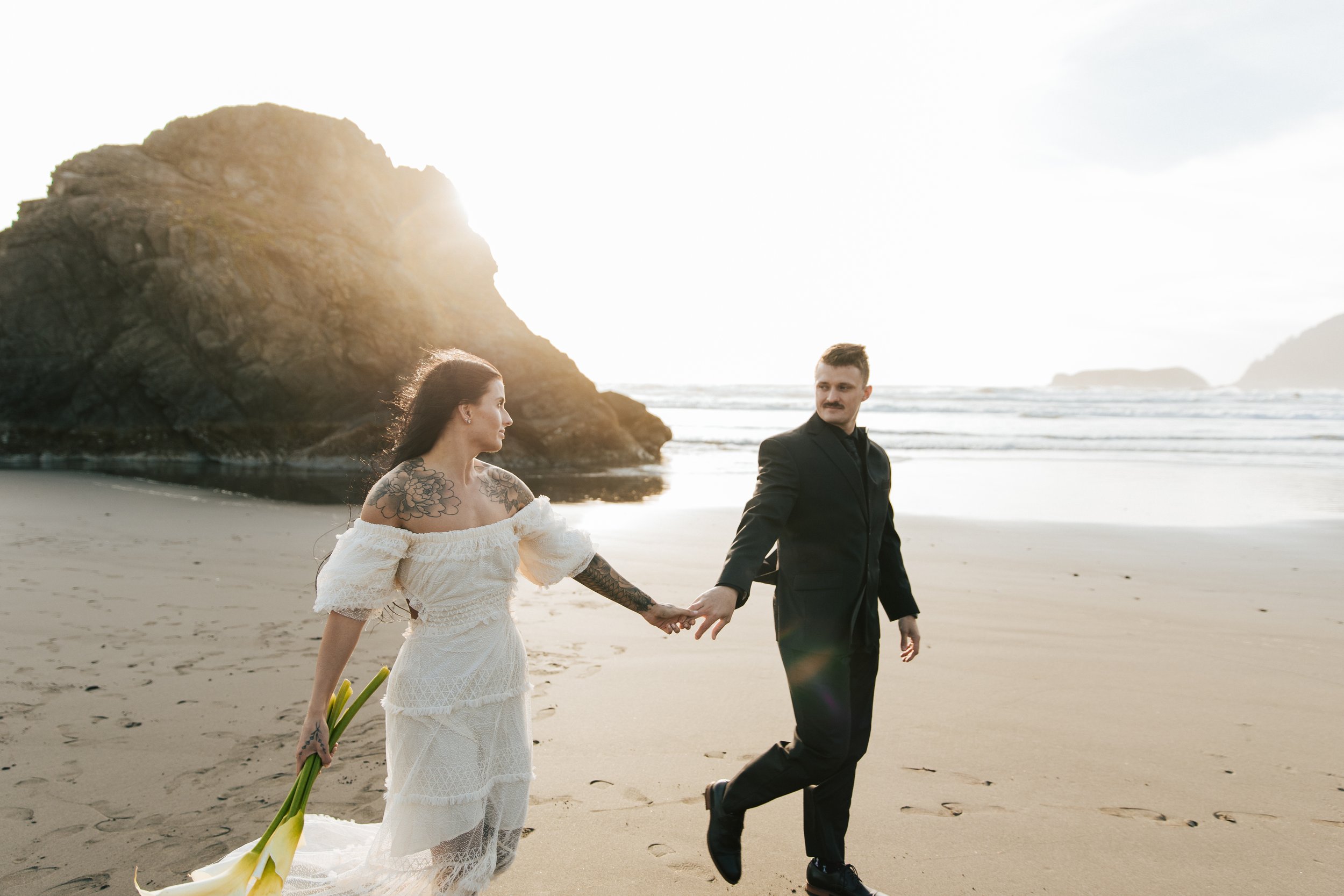  Oregon coast elopement. Couple elopes on the beach with the sun shining behind them. Sun rays behind rocks. Beach with rocks and haystacks. Southern Oregon. Couple walking together holding hands. Brookings, Oregon. Samuel H Boardman elopement. #oreg