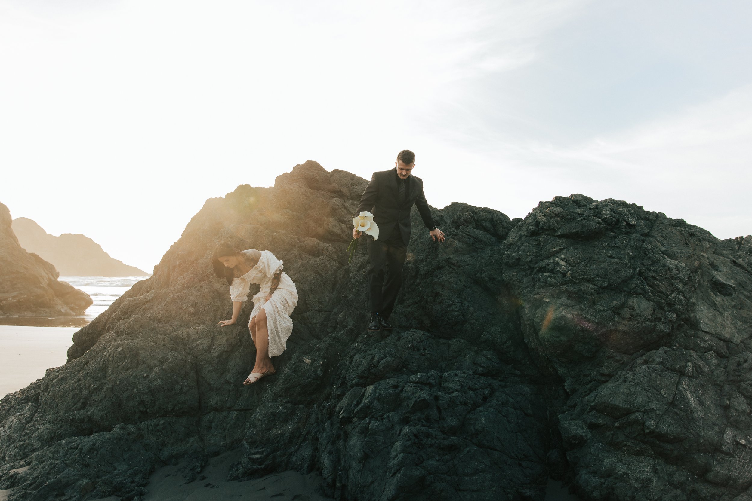  Oregon coast elopement. Couple elopes on the beach with the sun shining behind them. Sun rays behind rocks. Beach with rocks and haystacks. Southern Oregon. Couple climbs up large rock in wedding clothes. Brookings, Oregon. Samuel H Boardman elopeme