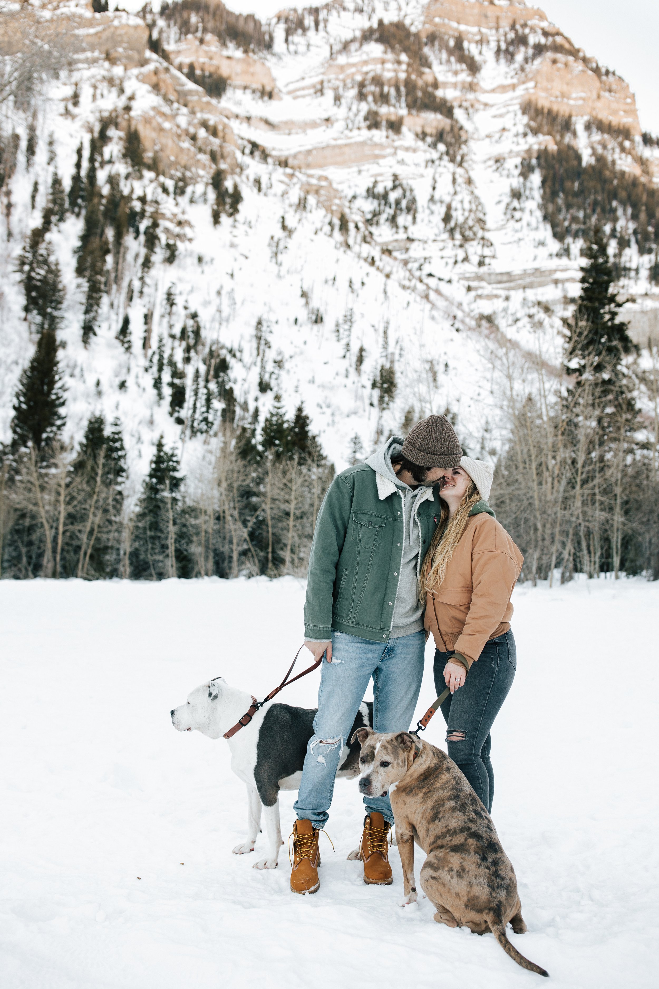  Winter photoshoot outfit inspo. Man and woman kiss in the snowy mountains wearing coats and beanies with their dogs. Utah mountain engagement shoot in the winter. Park City, Utah photographer. #winter #engagements #engagementsession #utahphotoshoot 