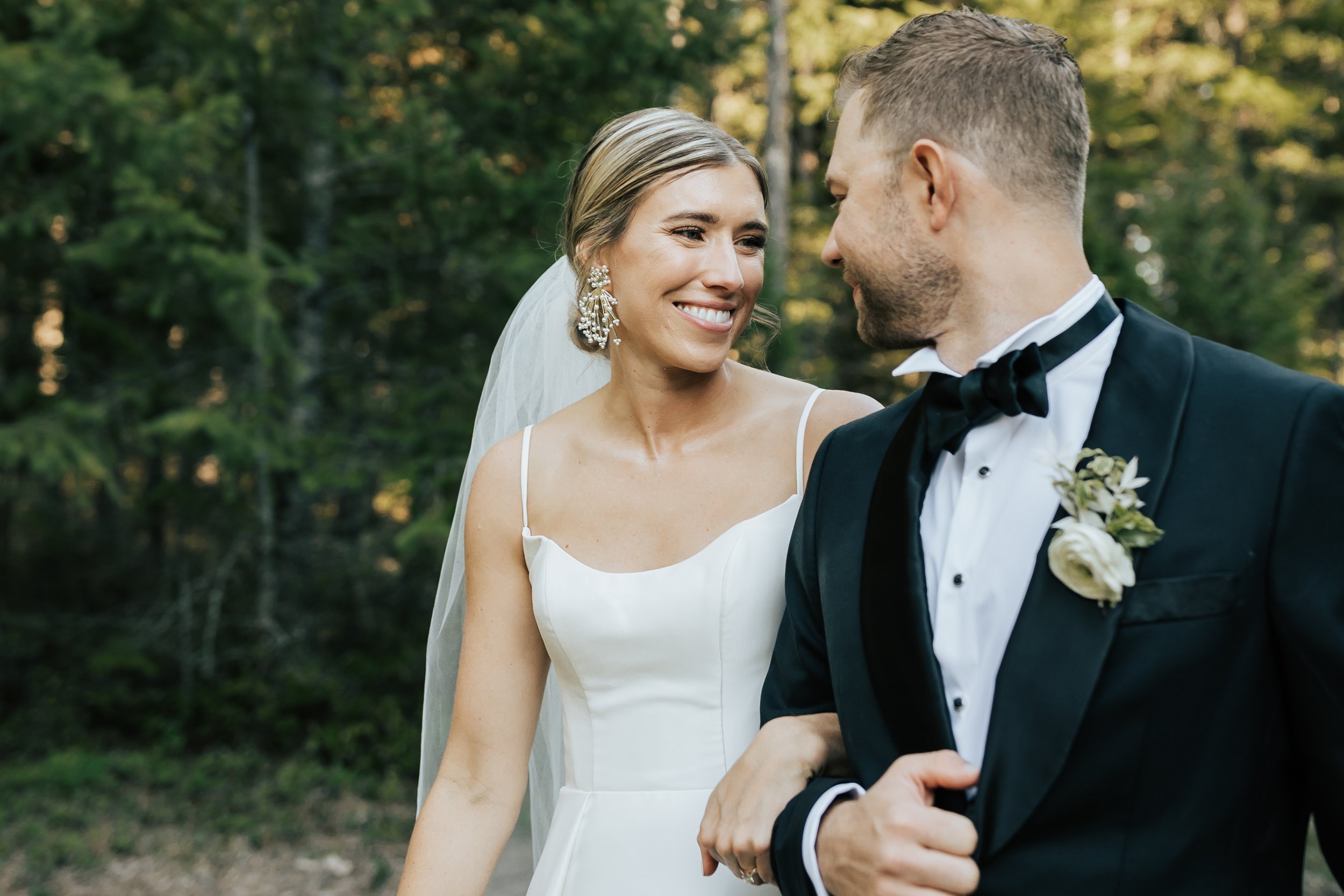  Newly married bride and groom smile at each other as they walk in the forest in Whitefish, Montana. Wedding inspo. Bride is wearing gorgeous large white earrings with a strappy wedding dress and groom is wearing a black suit and bowtie. #montanawedd