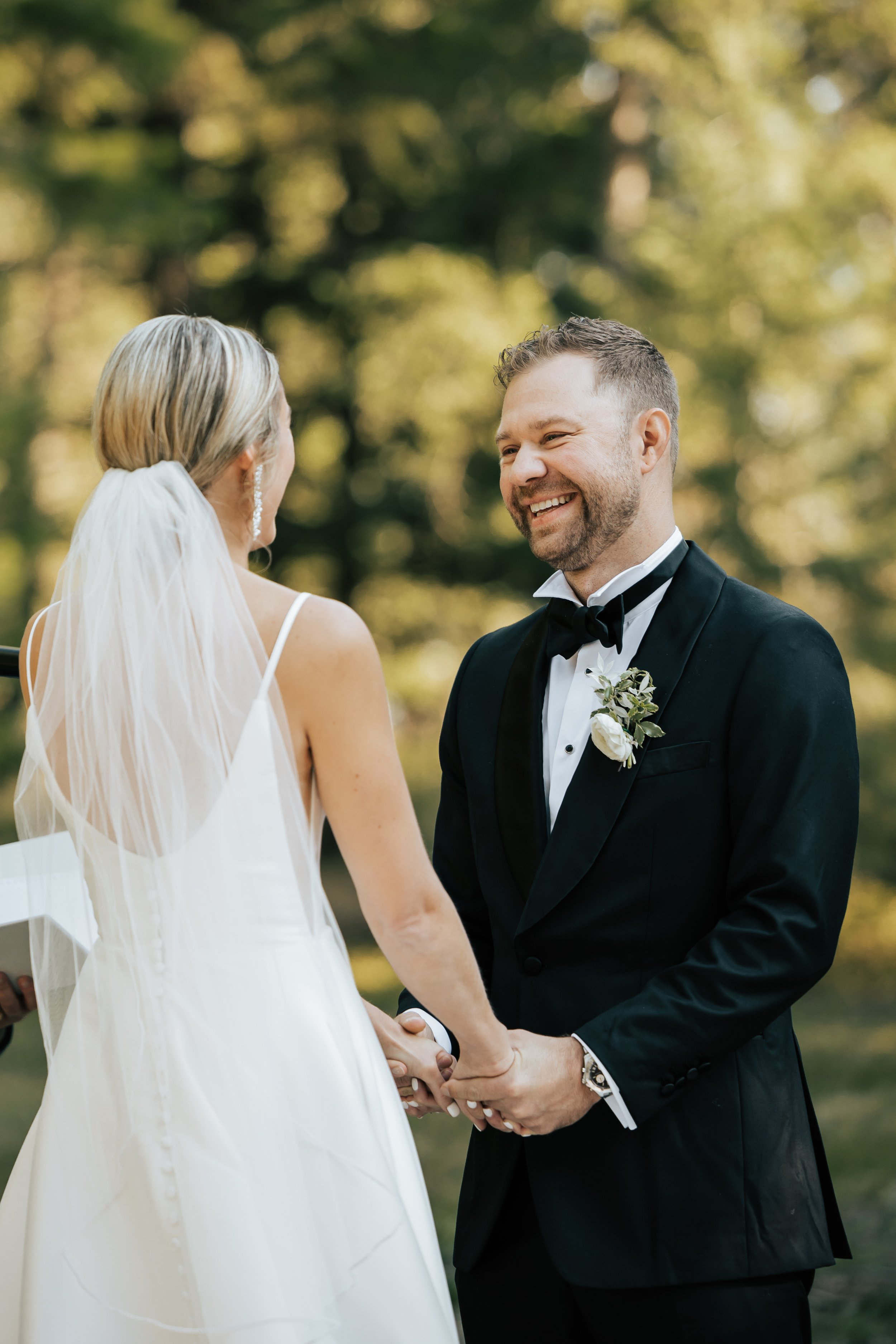  Bride and groom smile at each other during their wedding ceremony in the forest in Whitefish, Montana. Wedding inspo. Bride is wearing gorgeous large white earrings with a strappy wedding dress and veil and groom is wearing a black suit and bowtie. 