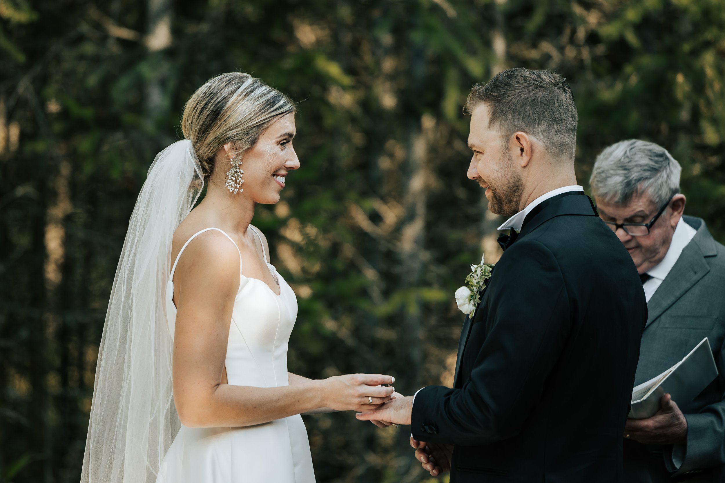  Bride and groom smile at each other during their wedding ceremony in the forest in Whitefish, Montana. Wedding inspo. Bride is wearing gorgeous large white earrings with a strappy wedding dress and veil and groom is wearing a black suit and bowtie. 