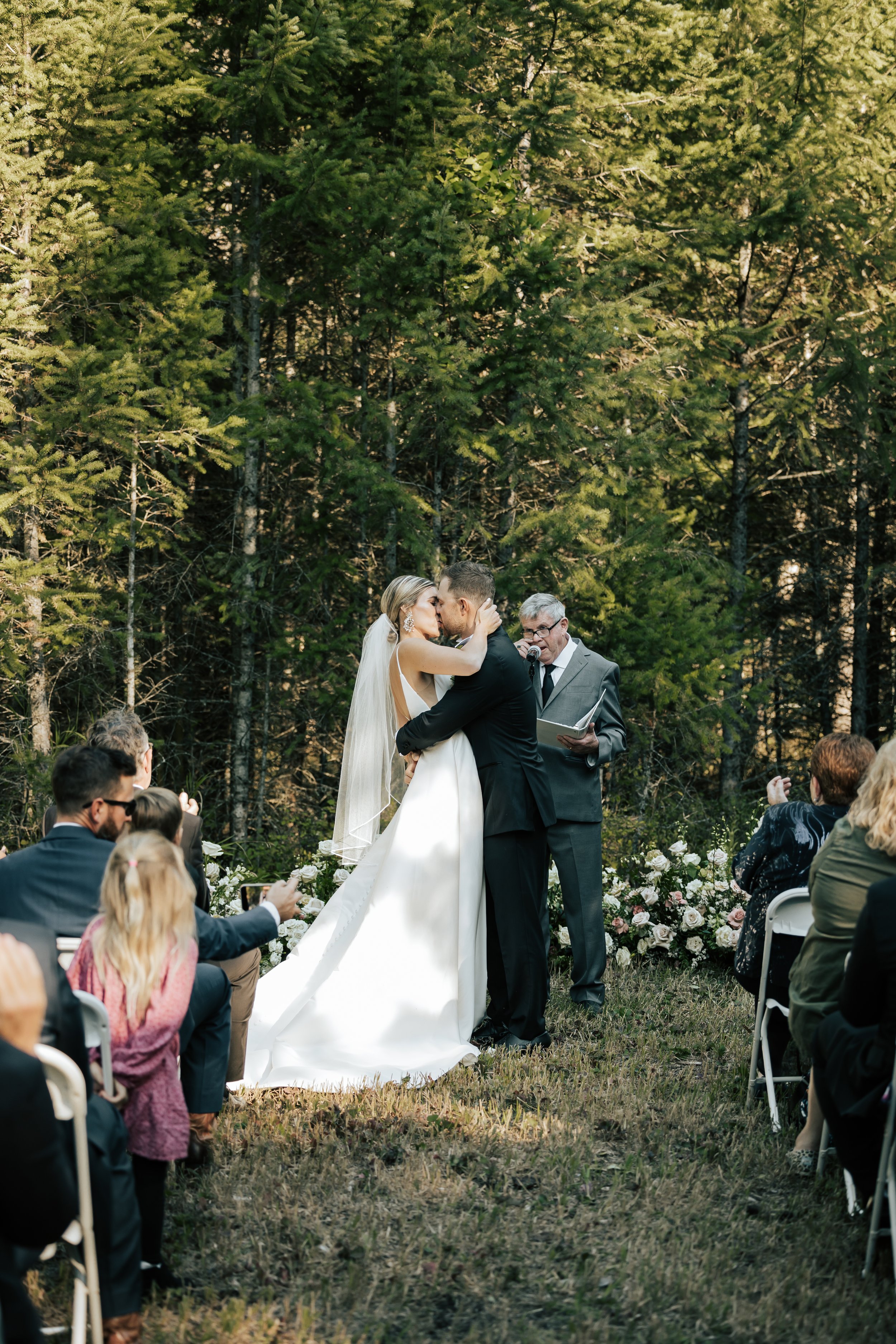  Bride and groom smile at each other during their wedding ceremony in the forest in Whitefish, Montana. Bride and groom kiss after they become husband and wife. Wedding inspo. Bride is wearing gorgeous large white earrings with a strappy wedding dres