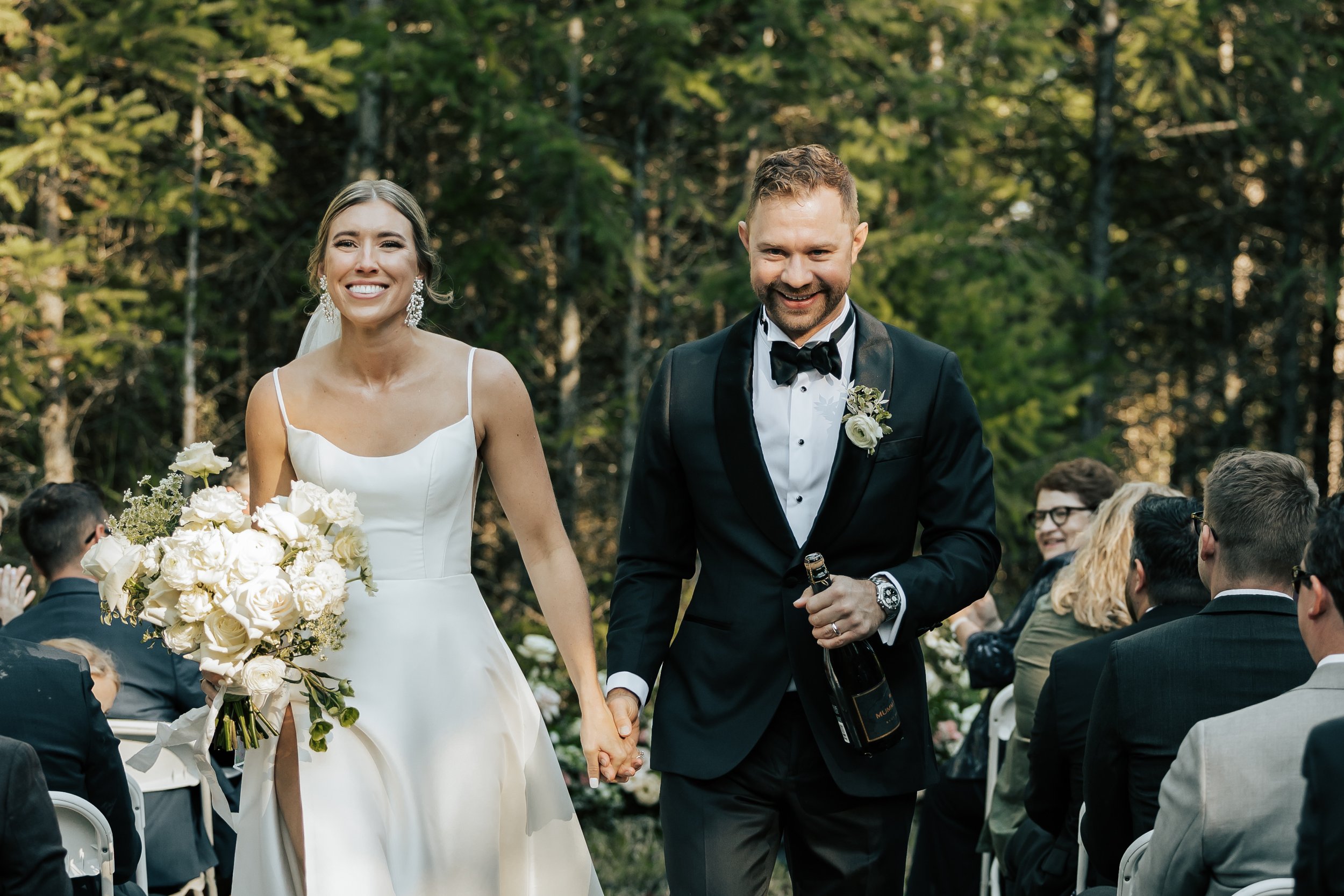  Bride and groom walk down the aisle together after getting married. Wedding ceremony in the forest in Whitefish, Montana. Wedding inspo. Bride is wearing gorgeous large white earrings with a strappy wedding dress and veil and groom is wearing a blac