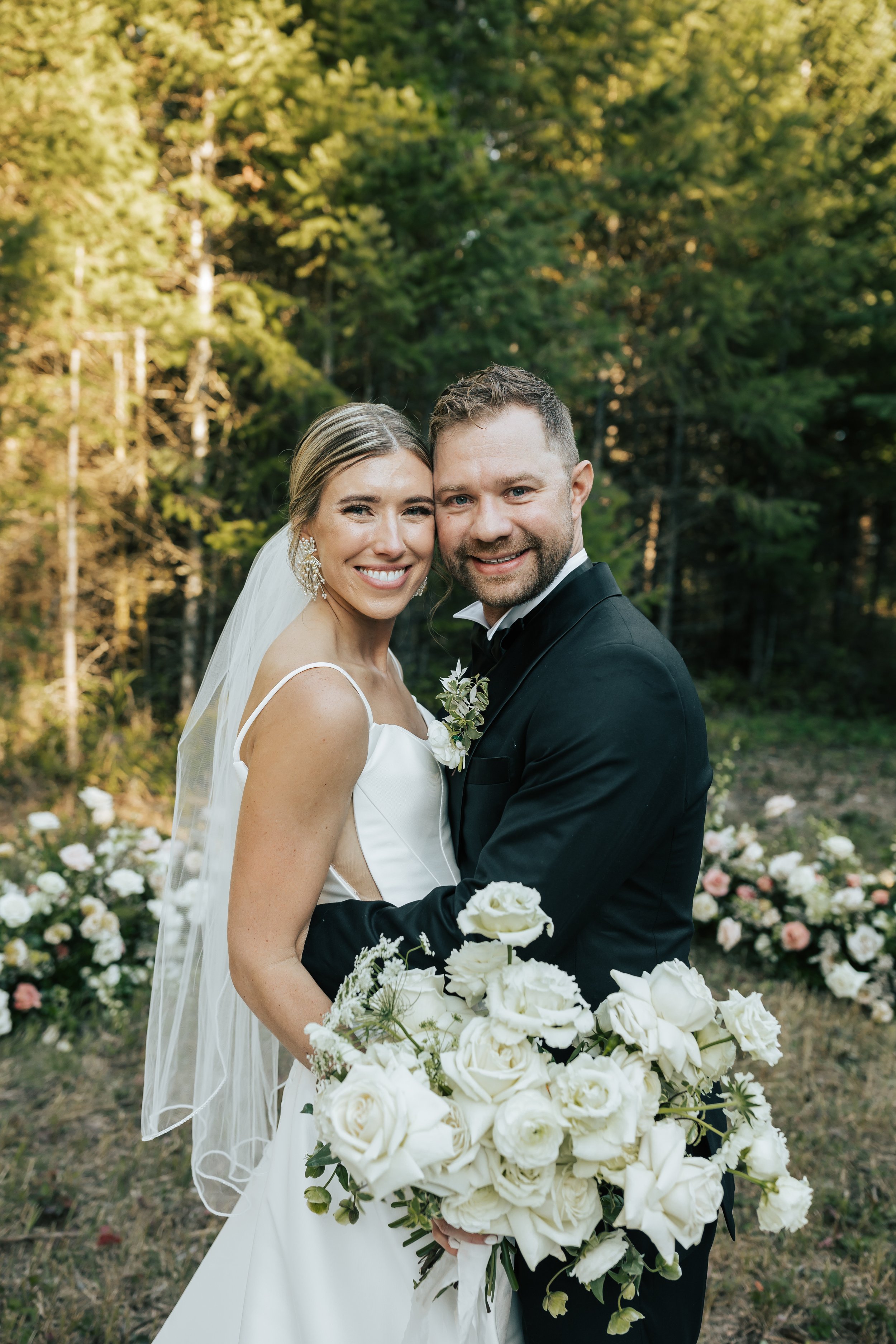  Bride and groom wedding portraits surrounded by beautiful flowers and pine trees. Bride and groom smile at wedding in the forest in Whitefish, Montana. Wedding inspo. Bride is wearing gorgeous large white earrings with a strappy wedding dress and ve