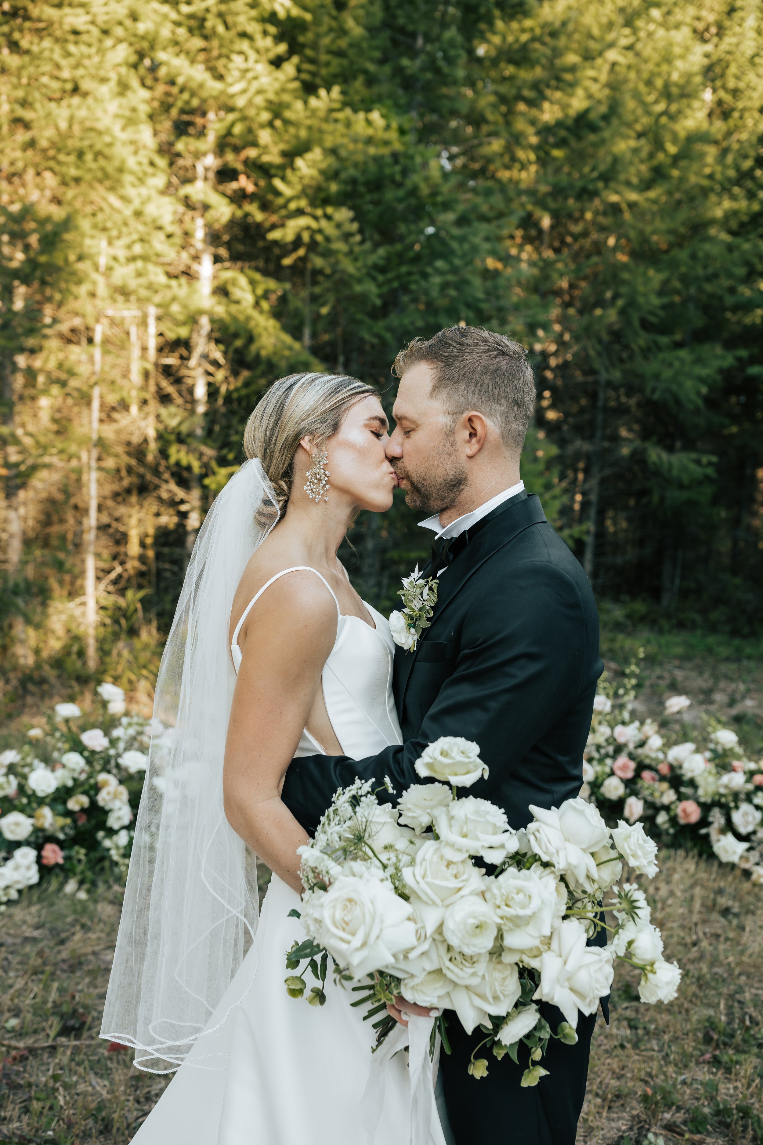  Bride and groom wedding portraits surrounded by beautiful flowers and pine trees. Bride and groom smile at wedding in the forest in Whitefish, Montana. Wedding inspo. Bride is wearing gorgeous large white earrings with a strappy wedding dress and ve