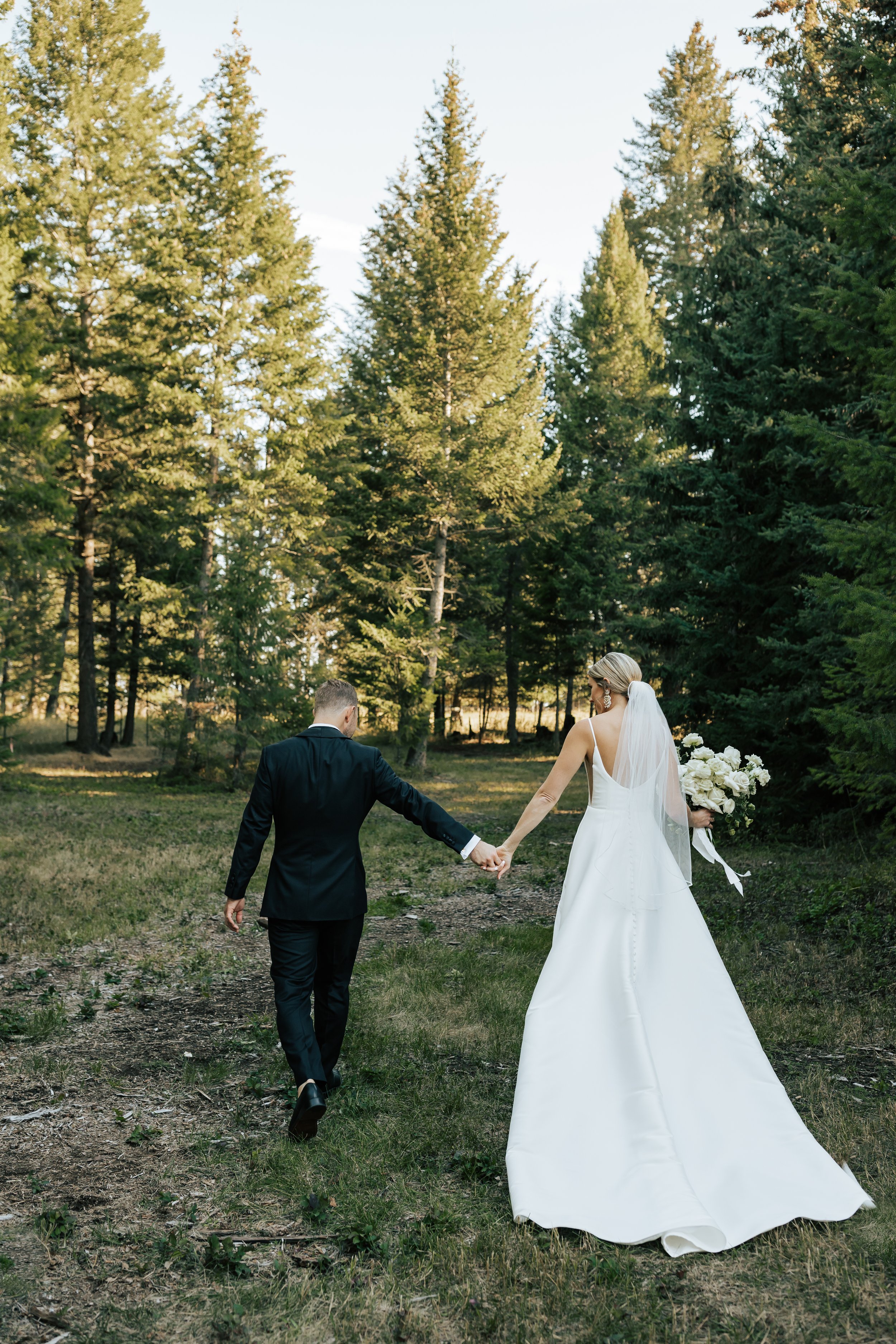  Bride and groom wedding portraits surrounded by beautiful flowers and pine trees. Bride and groom smile at wedding in the forest in Whitefish, Montana. Wedding inspo. Bride is wearing gorgeous large white earrings with a strappy wedding dress and ve