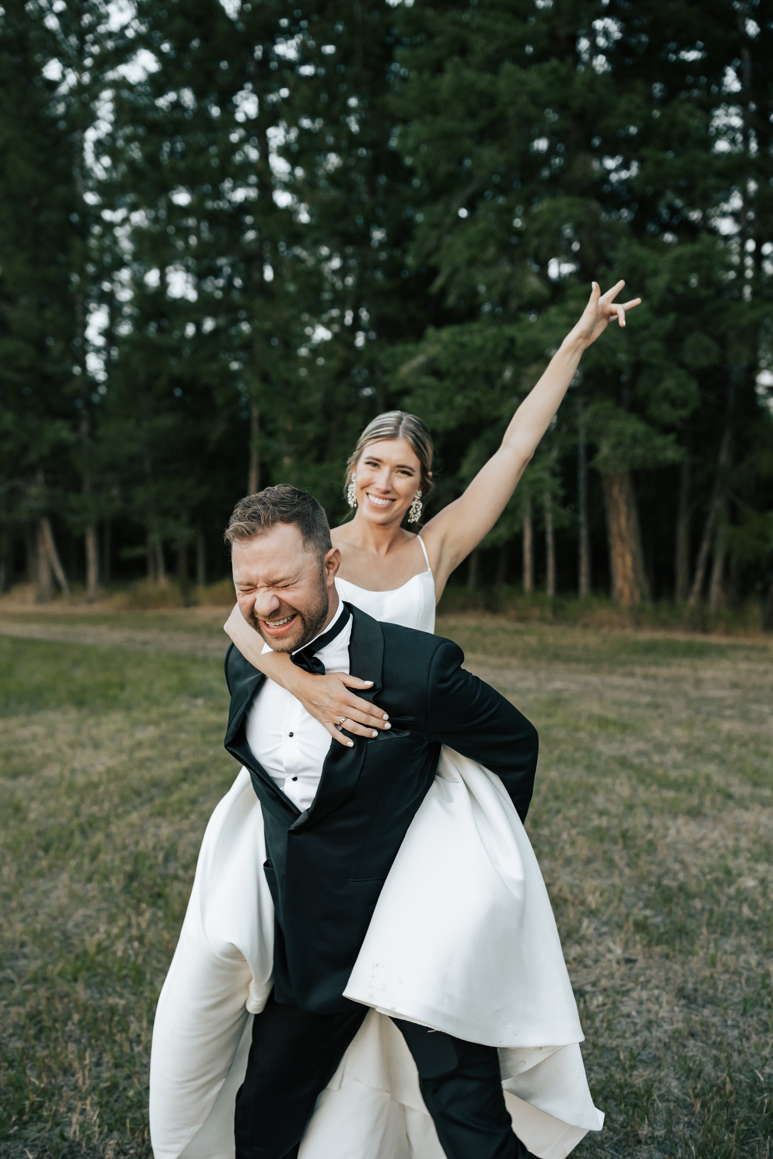  Bride and groom wedding portraits surrounded by beautiful flowers and pine trees. Bride and groom smile at wedding in the forest in Whitefish, Montana. Wedding inspo. Bride is wearing gorgeous large white earrings with a strappy wedding dress and ve
