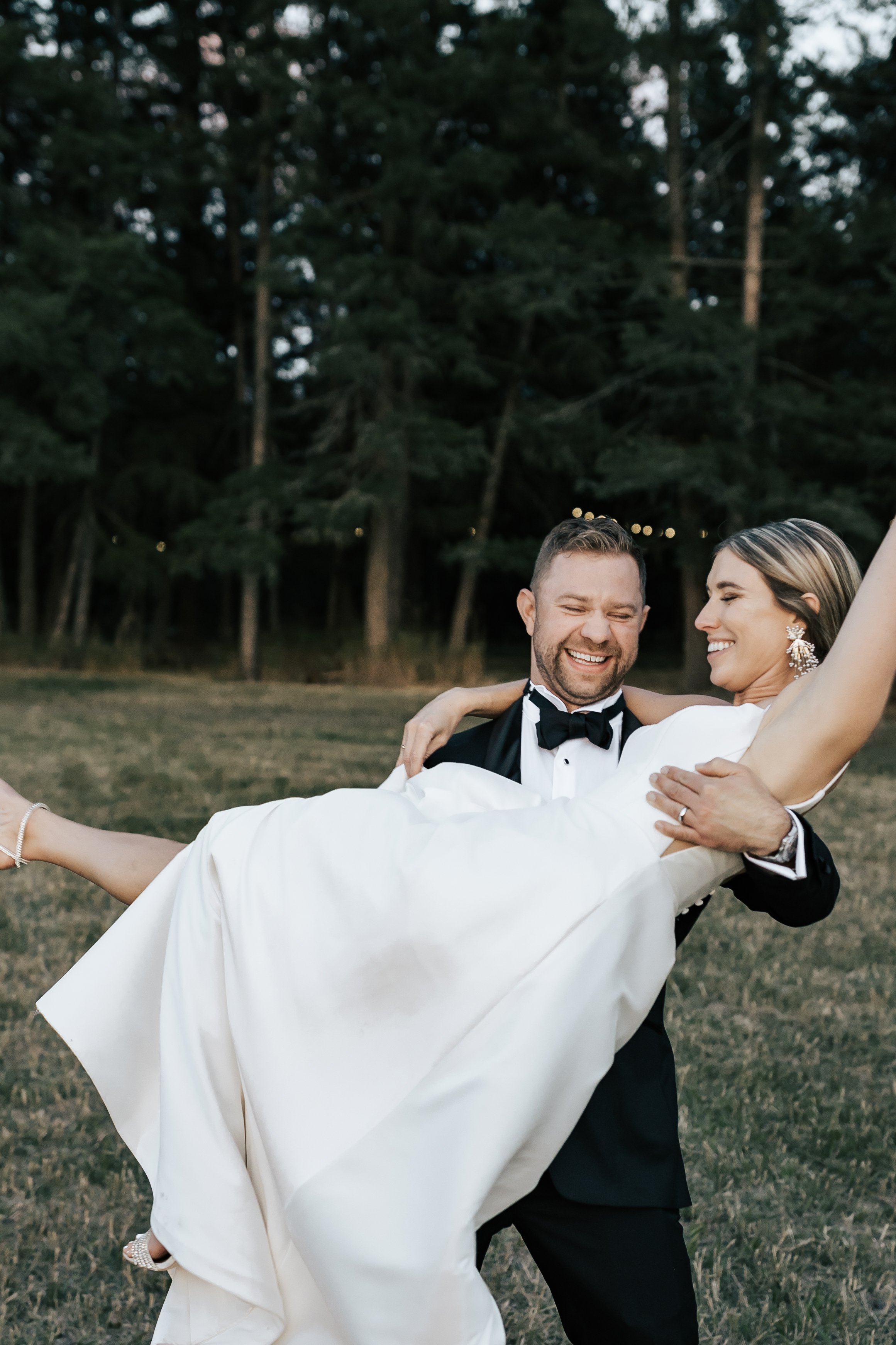  Bride and groom wedding portraits surrounded by pine trees. Bride and groom smile at wedding in the forest in Whitefish, Montana. Wedding inspo. Bride is wearing gorgeous large white earrings with a strappy wedding dress and veil and groom is wearin