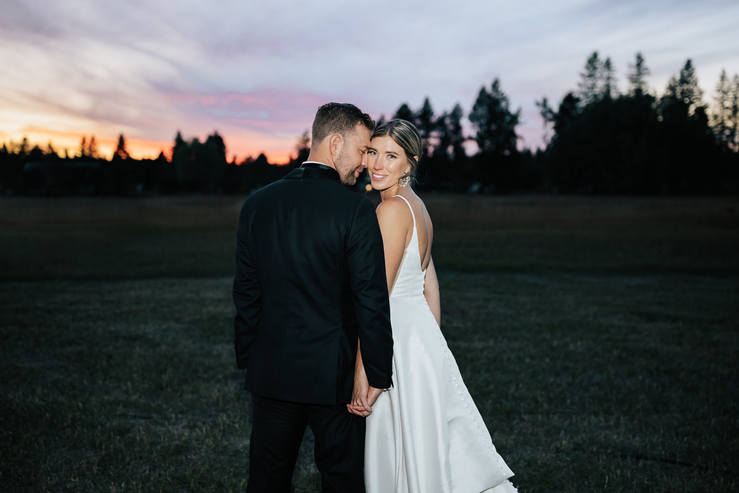  Bride and groom sunset wedding portraits surrounded by beautiful flowers and pine trees. Bride and groom smile at wedding in the forest in Whitefish, Montana. Wedding inspo. Bride is wearing gorgeous large white earrings with a strappy wedding dress