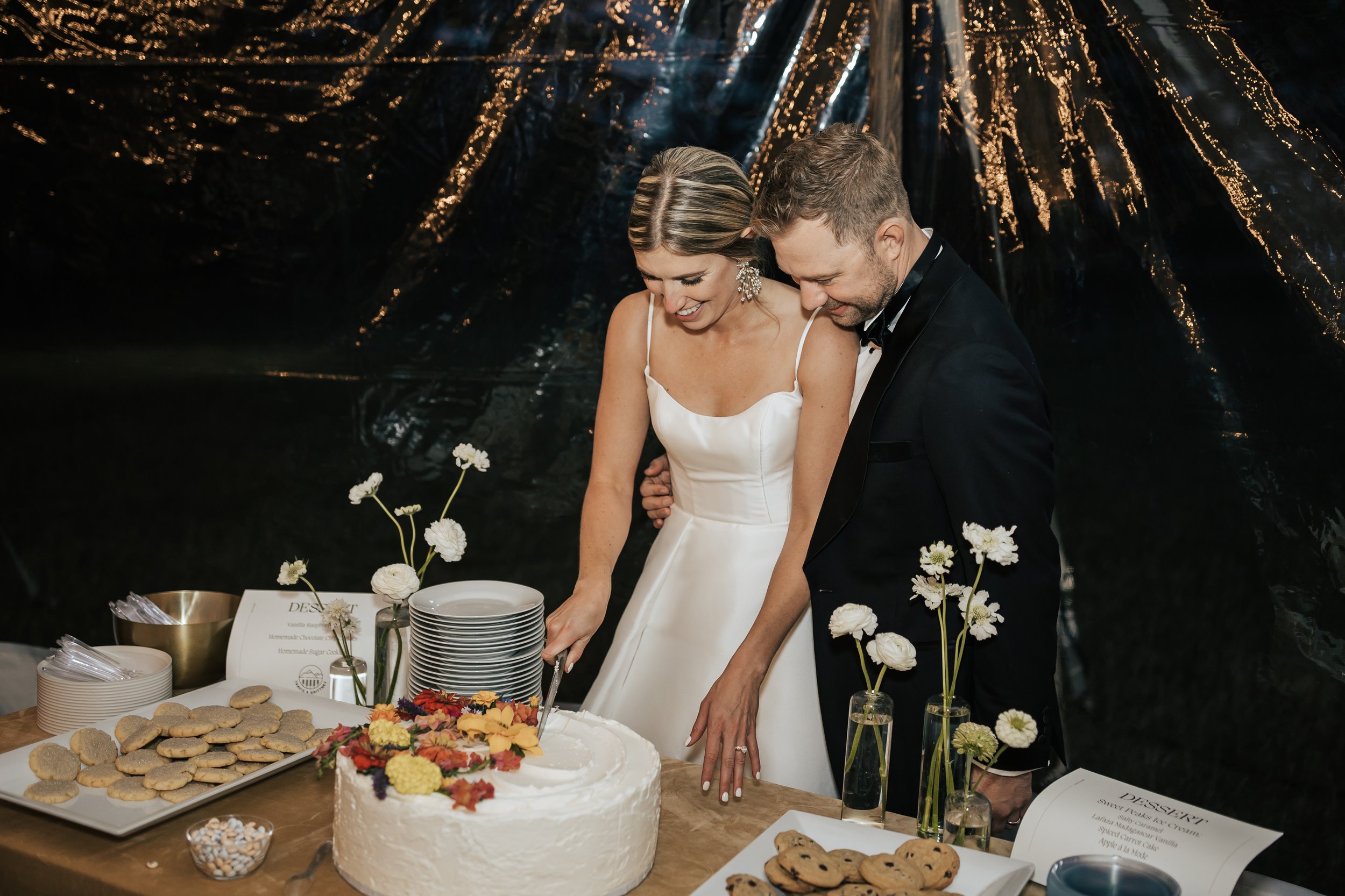  Bride and groom cutting cake. Wedding inspo. Bride is wearing gorgeous large white earrings with a strappy wedding dress and veil and groom is wearing a black suit and bowtie. #montanawedding #emilyjenkinsphoto #weddingphotos #summerwedding 