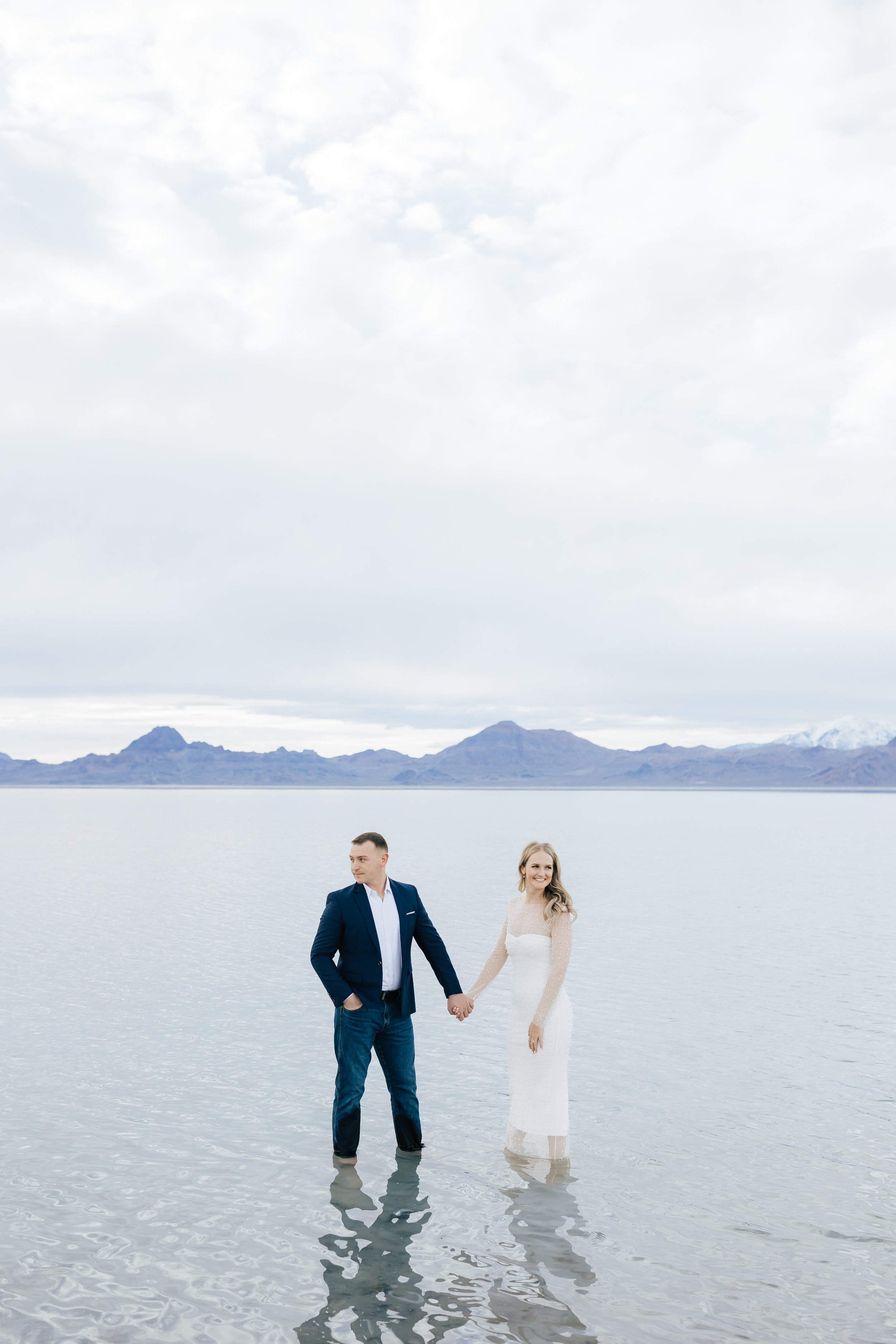  Emily Jenkins Photography captures a man and woman holding hands and walking through shallow water. blue suit and jeans engagement style ideas fancy engagement attire #EmilyJenkinsPhotography #EmilyJenkinsEngagements #BonnevilleSaltFlats #SaltFlatEn