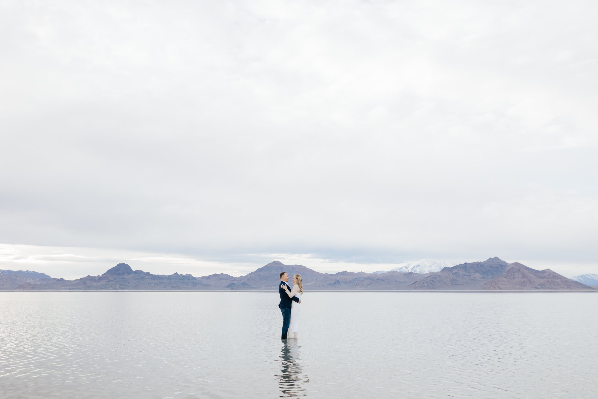  Emily Jenkins Photography captures a landscape view of a couple embracing at Bonneville Salt Flats. Stunning UT engagements shallow lake portraits #EmilyJenkinsPhotography #EmilyJenkinsEngagements #BonnevilleSaltFlats #SaltFlatEngagements #UtahEngag