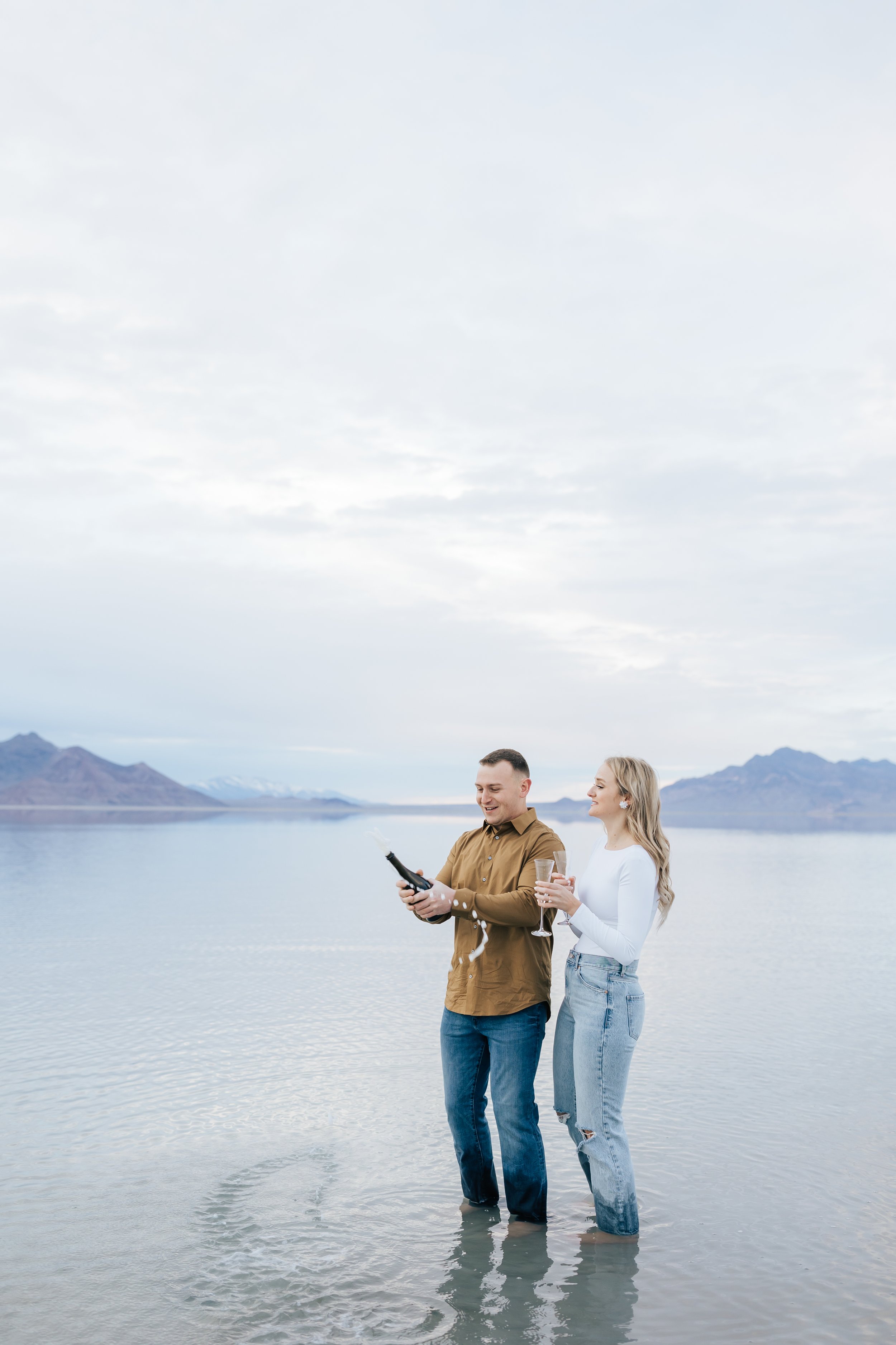  A man in a camel-colored button-up pops the bottle of champagne to celebrate his engagement with Emily Jenkins Photography. popp the bottle button up shirt celebrating couple #EmilyJenkinsPhotography #EmilyJenkinsEngagements #BonnevilleSaltFlats #Sa