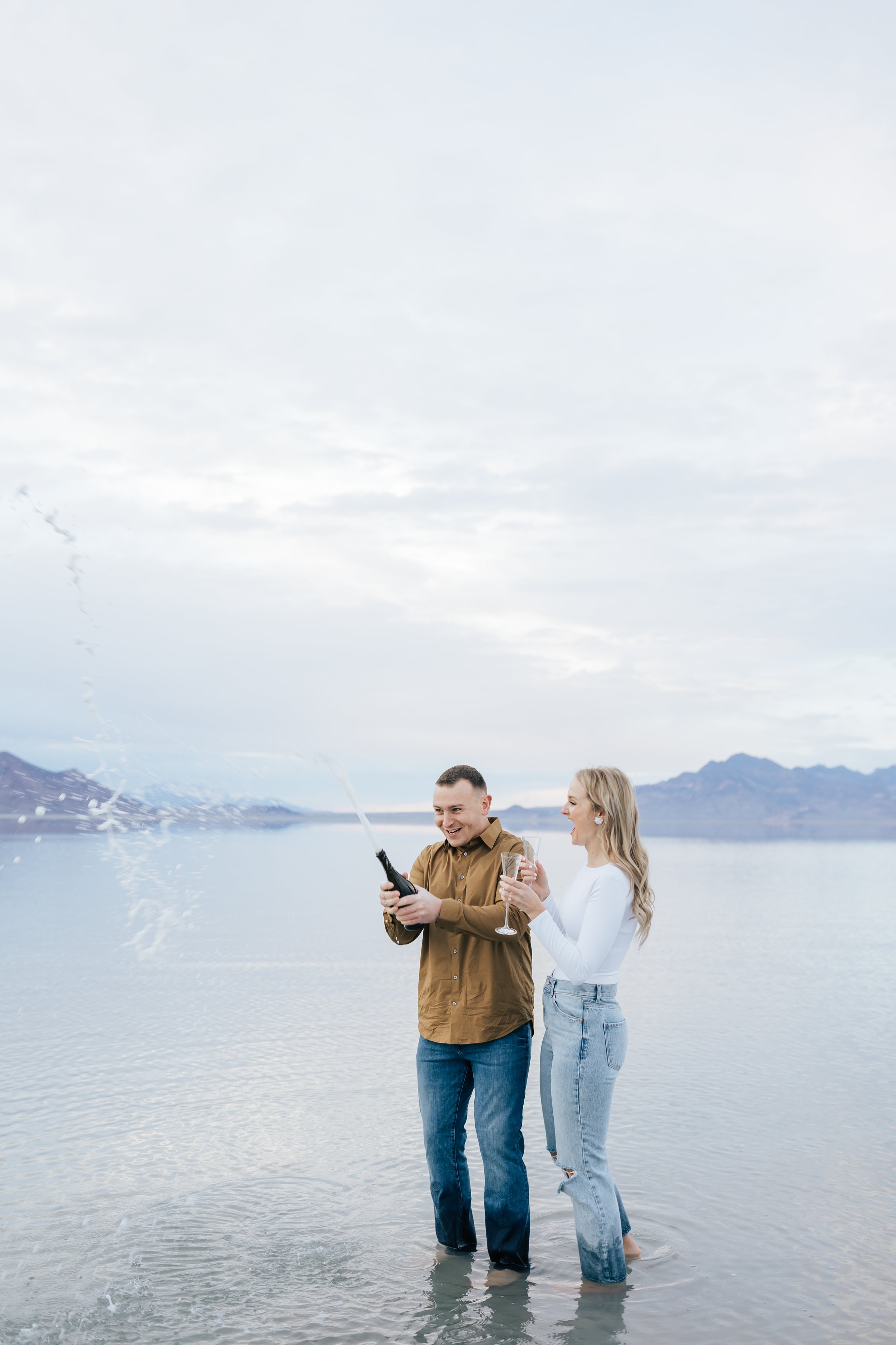  An engaged couple laughs as the champagne bottle sprays everywhere captured by Emily Jenkins Photography in Bonneville. Bonneville Engagements Utah Bonneville Photography #EmilyJenkinsPhotography #EmilyJenkinsEngagements #BonnevilleSaltFlats #SaltFl