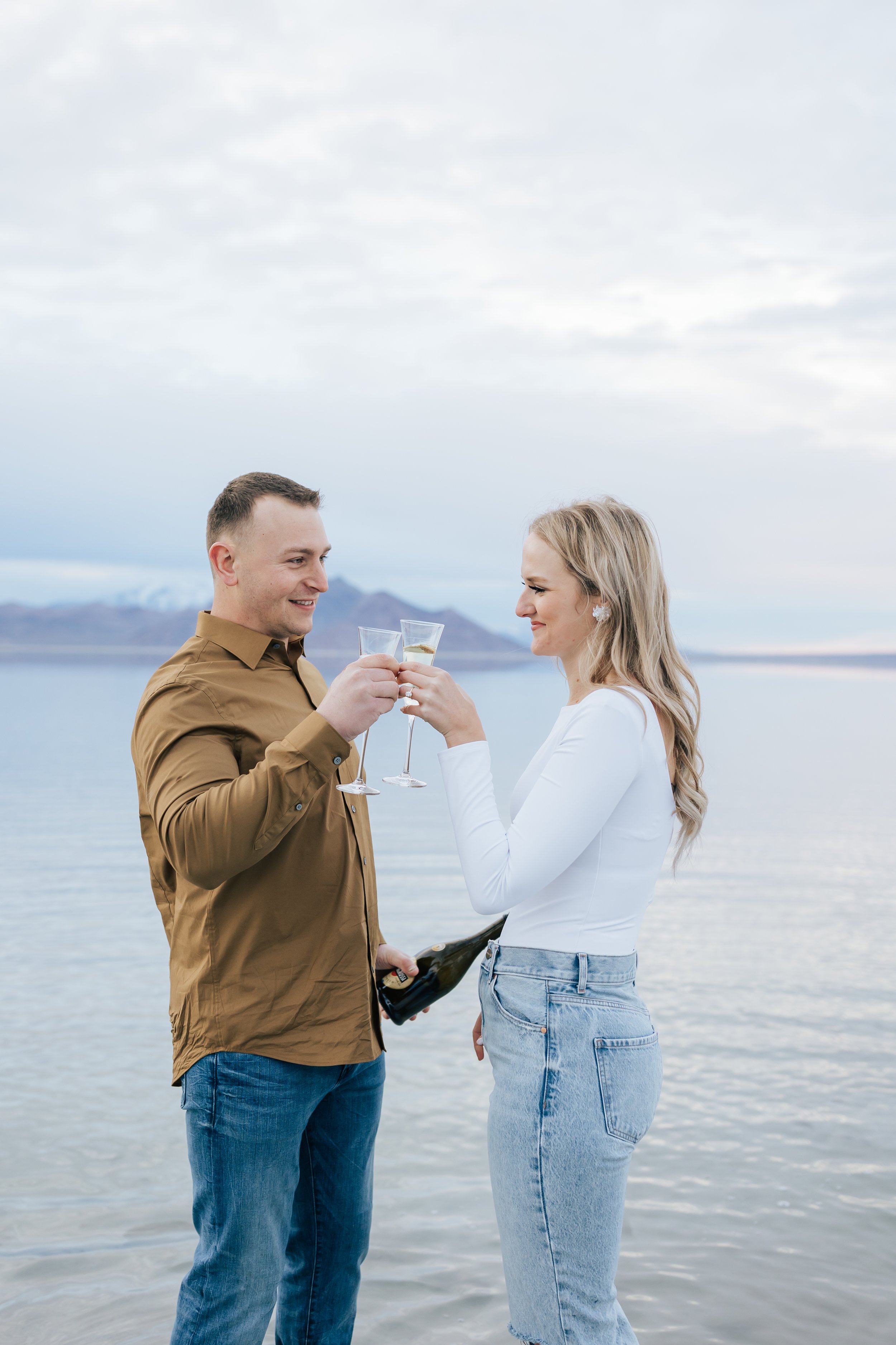  A woman in jeans and a white shirt clinks her glass with her boyfriend celebrating their engagement by Emily Jenkins Photography. clink the champagne glasses #EmilyJenkinsPhotography #EmilyJenkinsEngagements #BonnevilleSaltFlats #SaltFlatEngagements