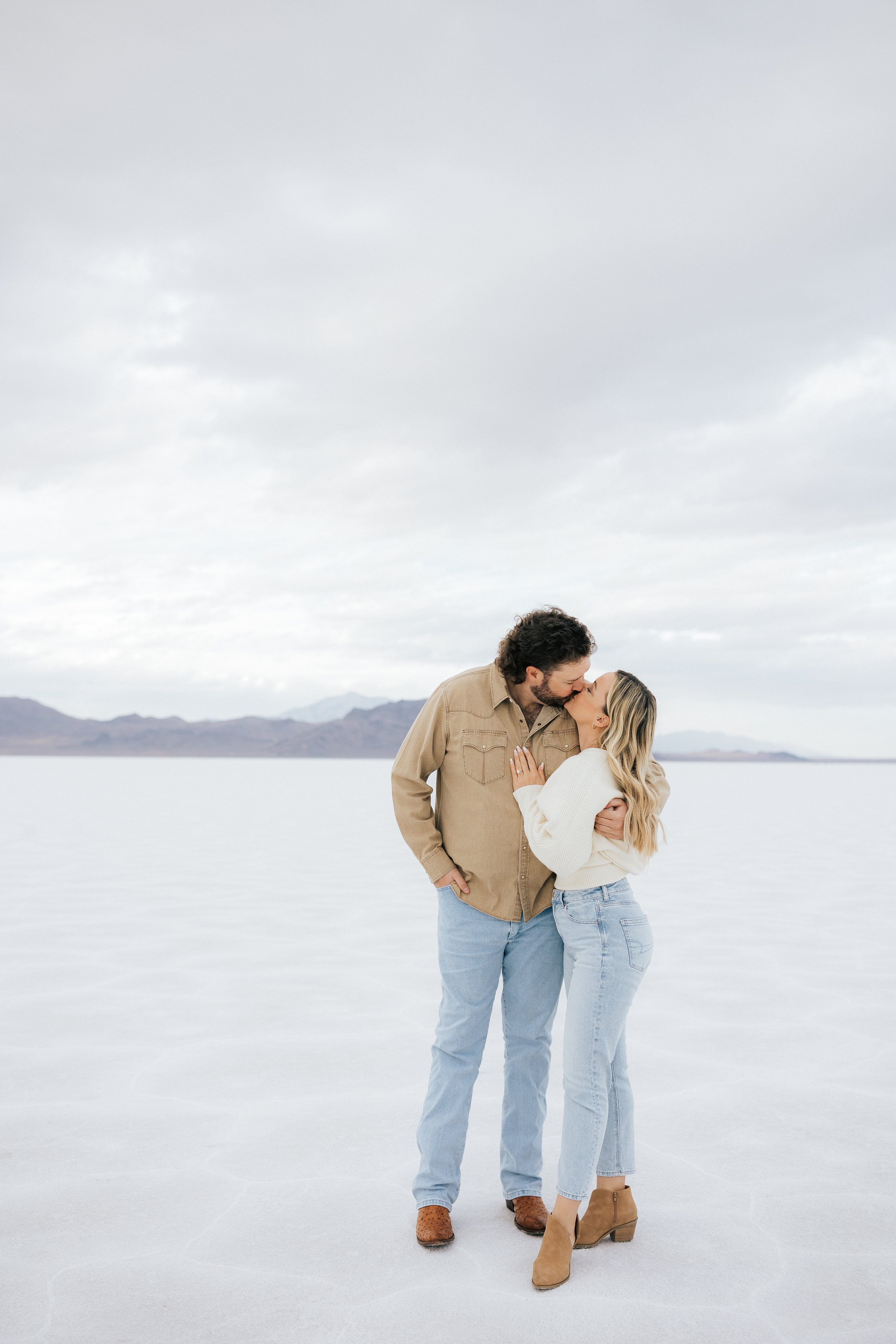  An engaged couple kisses at the Bonneville Salt Flats near Wendover, Nevada and Salt Lake City, Utah. The salt flats are overcast and white with cracks and mountains behind them. The girl wears a white sweater with jeans and the man wears a tan butt