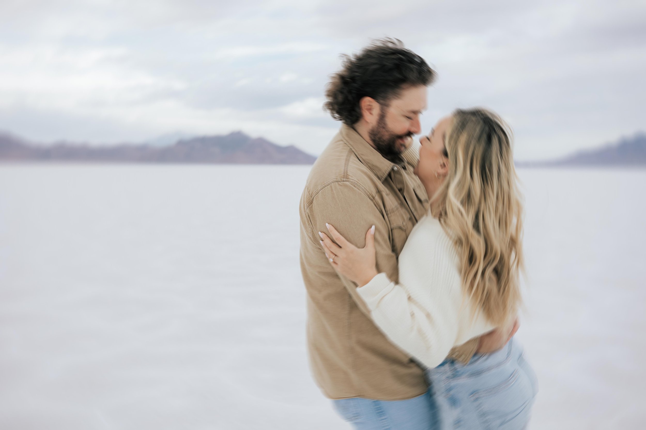  An engaged couple laughs and hugs as they pose for a blurry photo at the Bonneville Salt Flats near Wendover, Nevada and Salt Lake City, Utah. The Salt Flats are white and the sky is overcast. The mountains show behind. Engagement session at the Uta