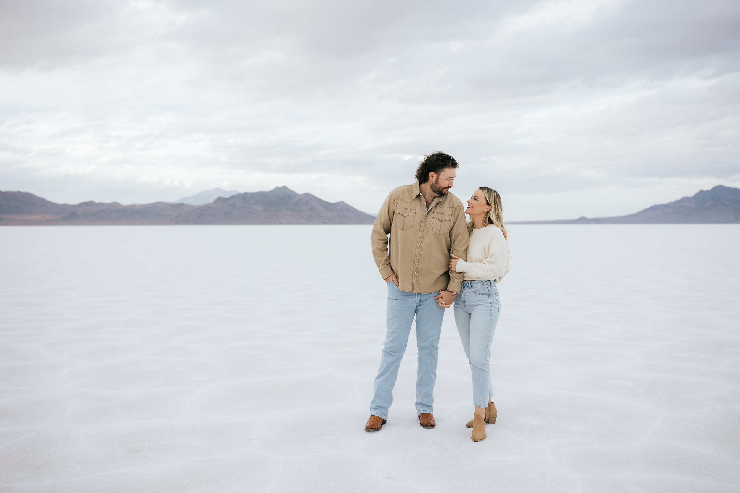  An engaged couple walks together while holding hands as they pose for a photo at the Bonneville Salt Flats near Wendover, Nevada and Salt Lake City, Utah. The Salt Flats are white and the sky is overcast. The mountains show behind. Engagement sessio