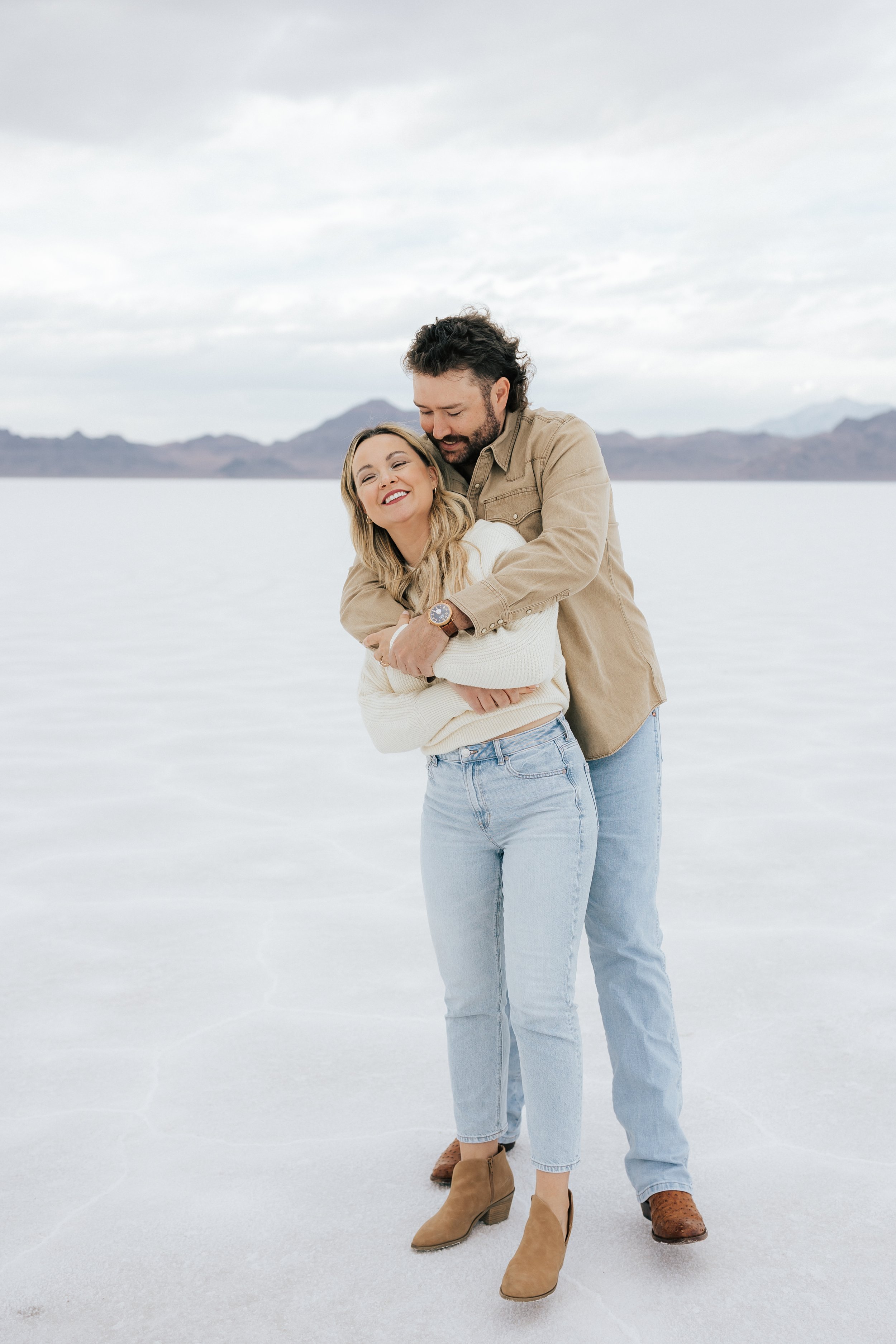  An engaged couple laughs and hugs as they pose for a photo at the Bonneville Salt Flats near Wendover, Nevada and Salt Lake City, Utah. The Salt Flats are white and the sky is overcast. The mountains show behind. Engagement session at the Utah Salt 
