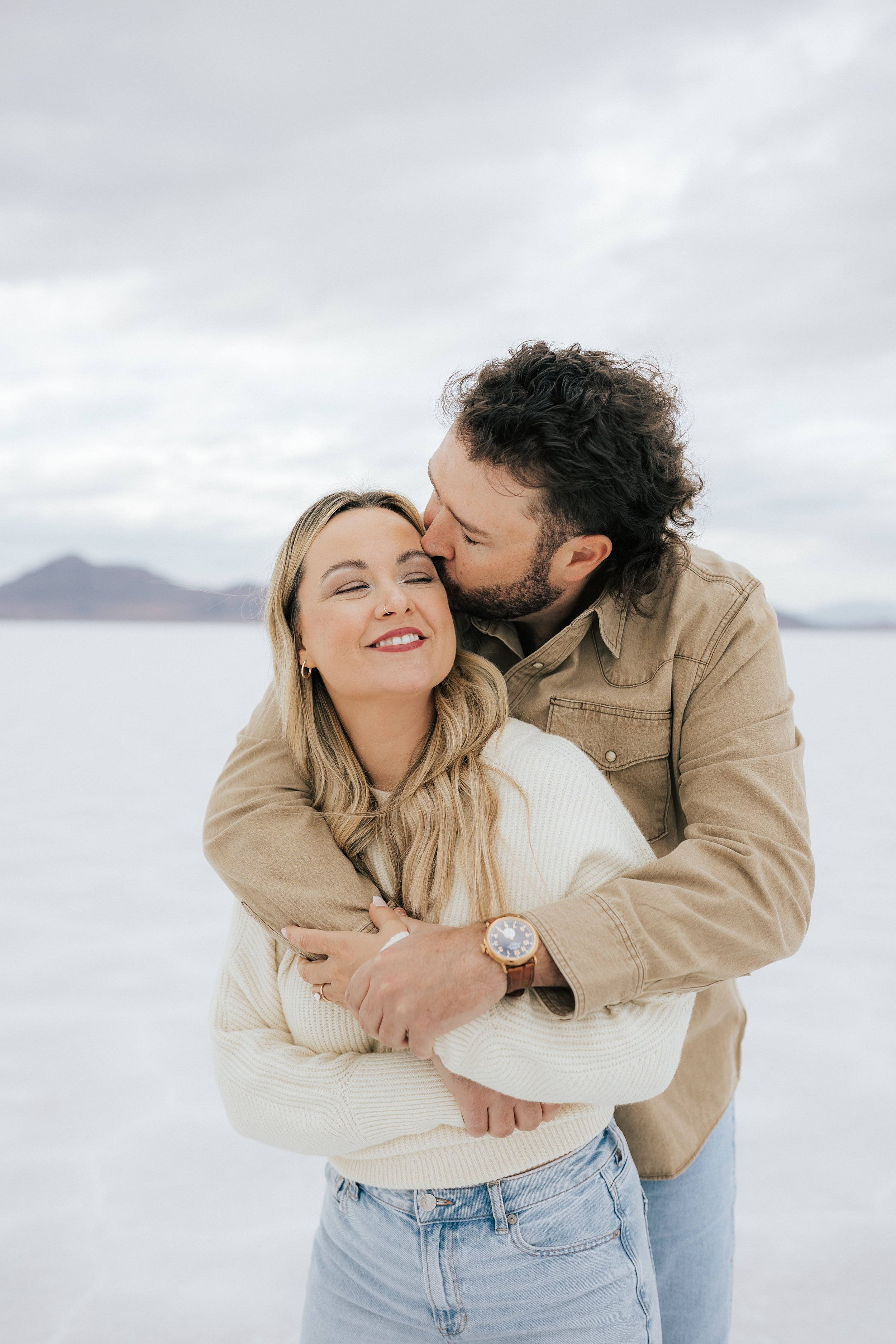  An engaged couple laughs and hugs as they pose for a photo at the Bonneville Salt Flats near Wendover, Nevada and Salt Lake City, Utah. The Salt Flats are white and the sky is overcast. The mountains show behind. Engagement session at the Utah Salt 