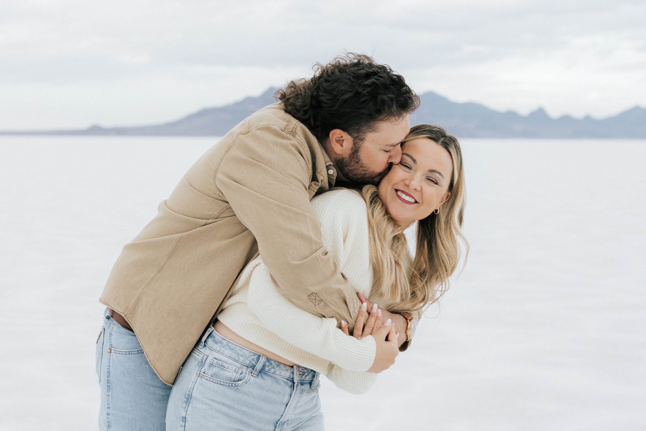  An engaged couple laughs and hugs as they pose for a photo at the Bonneville Salt Flats near Wendover, Nevada and Salt Lake City, Utah. Man kisses his fiance on the cheek. The Salt Flats are white and the sky is overcast. The mountains show behind. 