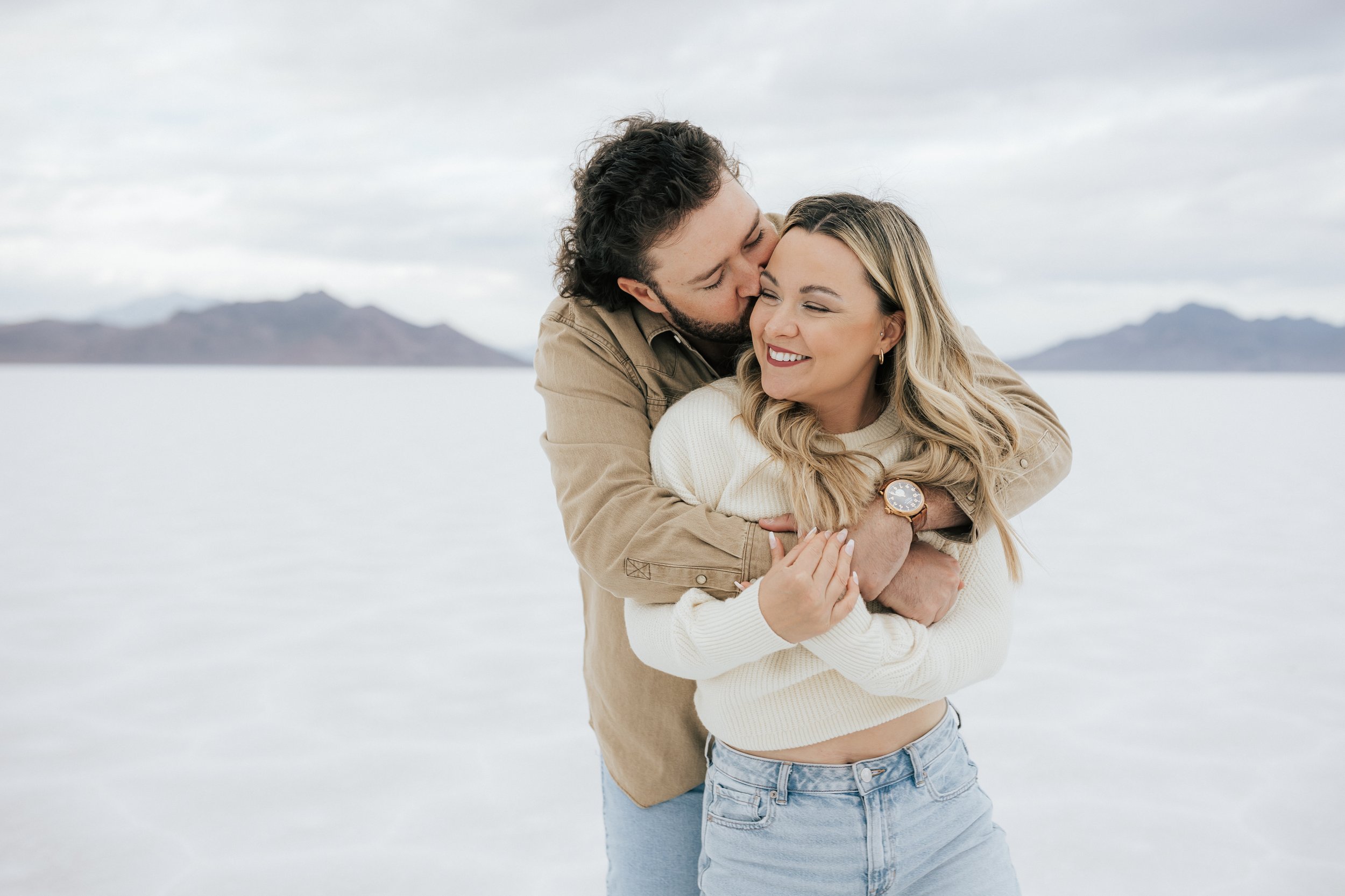  An engaged couple laughs and hugs as they pose for a photo at the Bonneville Salt Flats near Wendover, Nevada and Salt Lake City, Utah. Man kisses fiance on the cheek. The Salt Flats are white and the sky is overcast. The mountains show behind. Enga