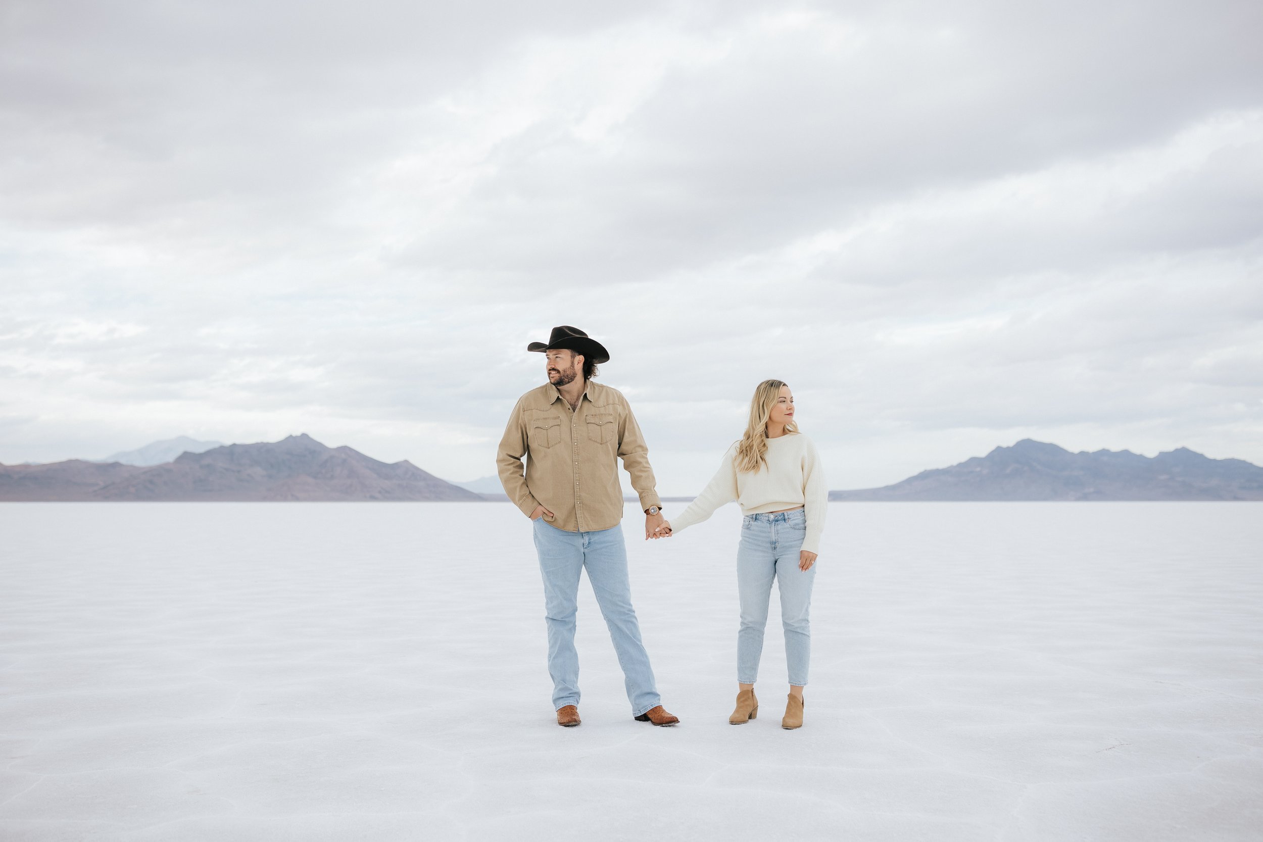  An engaged couple pose for a photo at the Bonneville Salt Flats near Wendover, Nevada and Salt Lake City, Utah. The guy wears a cowboy hat and the couple is looking away from each other. The Salt Flats are white and the sky is overcast. The mountain