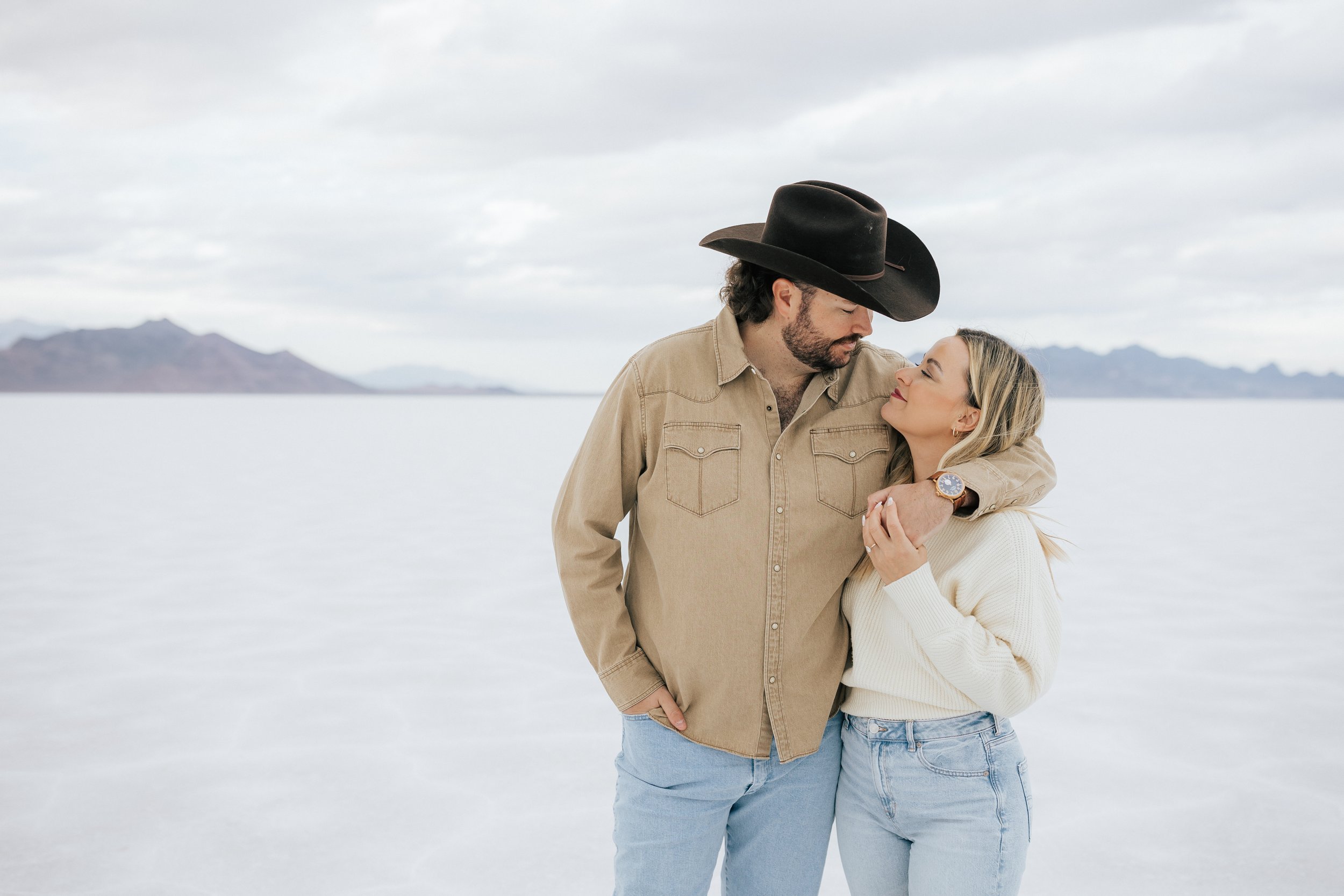 An engaged couple laughs and hugs as they pose for a photo at the Bonneville Salt Flats near Wendover, Nevada and Salt Lake City, Utah. The man is wearing a cowboy hat as they look at each other. The Salt Flats are white and the sky is overcast. The