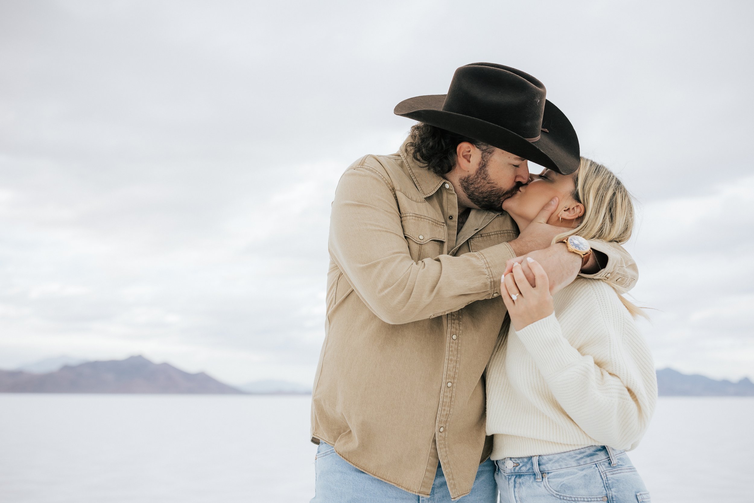  An engaged couple kisses at the Bonneville Salt Flats near Wendover, Nevada and Salt Lake City, Utah. The man is holding his girl’s neck as he kisses her. The Salt Flats are white and the sky is overcast. The mountains show behind. Engagement sessio