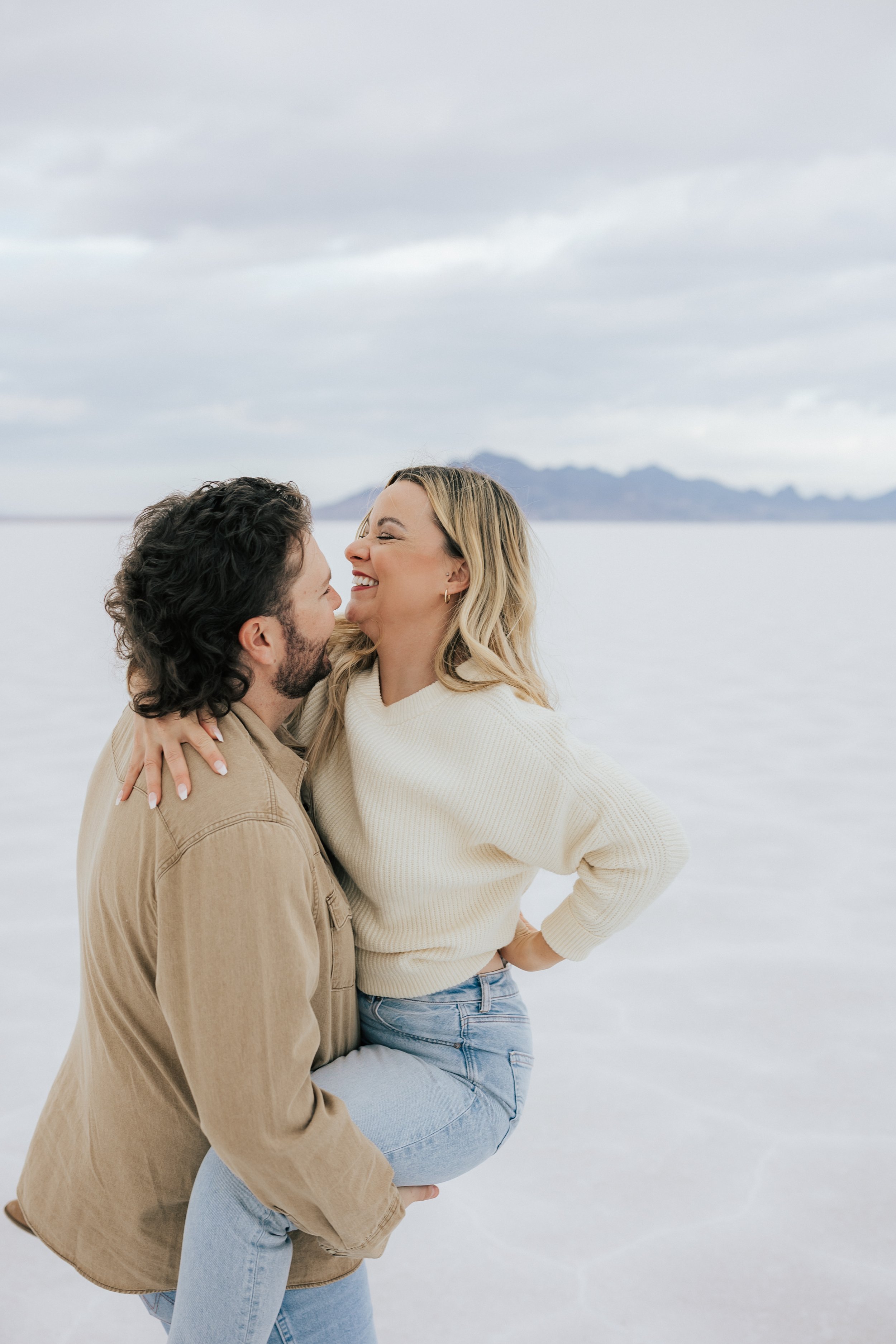  An engaged couple laughs and hugs as the man picks up and carries his girl for a photo at the Bonneville Salt Flats near Wendover, Nevada and Salt Lake City, Utah. The Salt Flats are white and the sky is overcast. The mountains show behind. Engageme