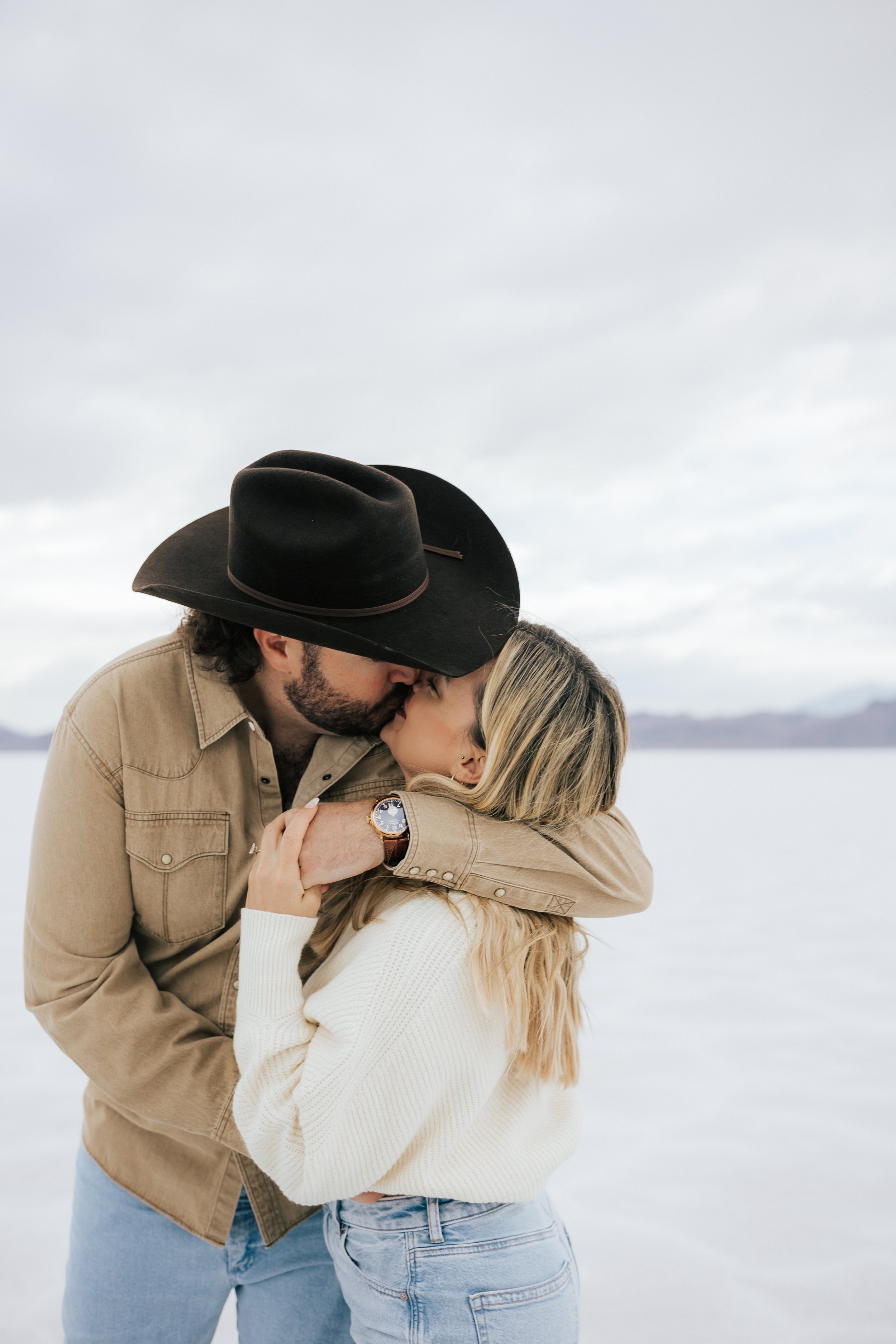  An engaged couple kisses and hugs as they pose for a photo at the Bonneville Salt Flats near Wendover, Nevada and Salt Lake City, Utah. The Salt Flats are white and the sky is overcast. The mountains show behind. Engagement session at the Utah Salt 