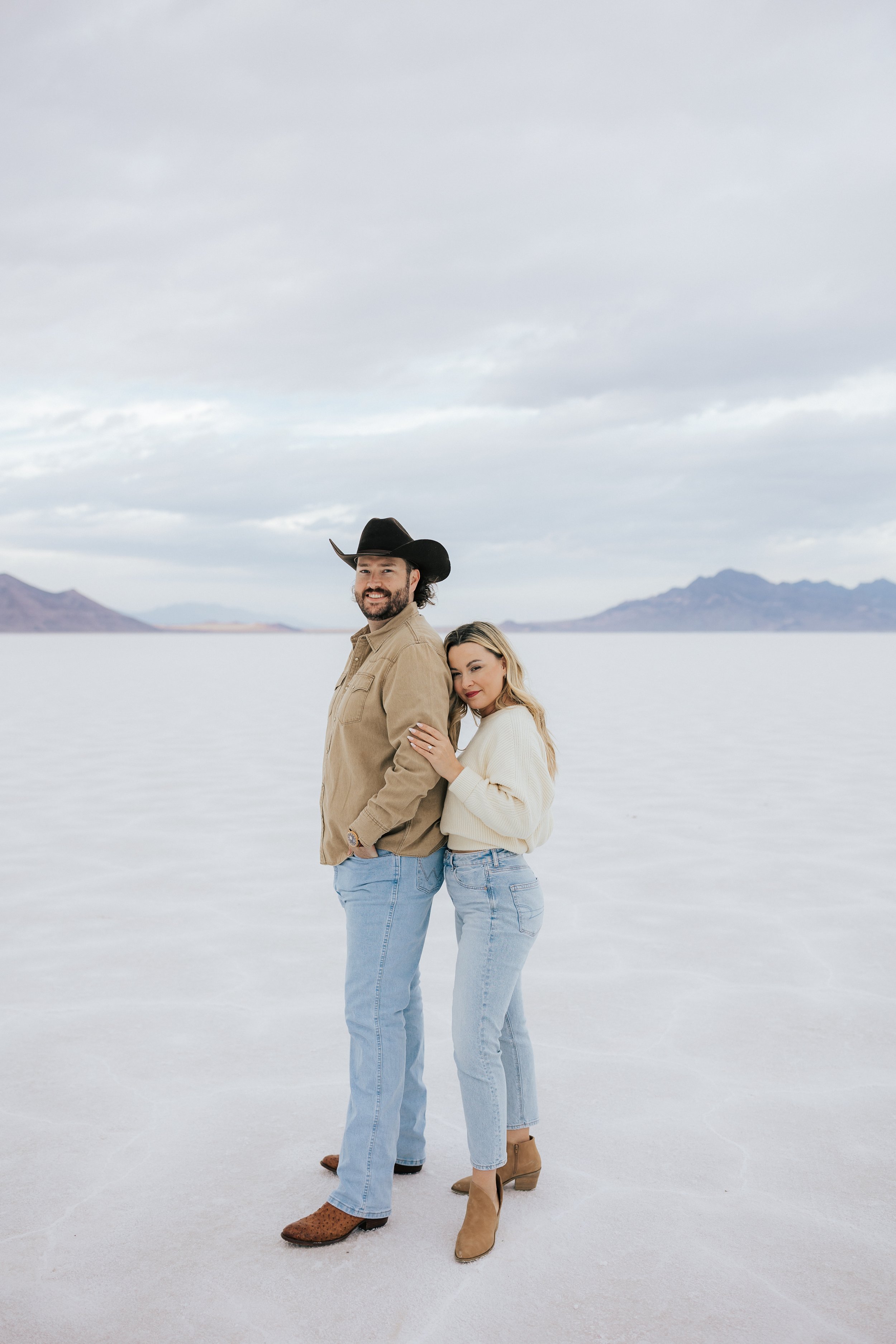  An engaged couple poses for a photo at the Bonneville Salt Flats near Wendover, Nevada and Salt Lake City, Utah. The Salt Flats are white and the sky is overcast. The mountains show behind. Engagement session at the Utah Salt Flats. Salt Flats photo