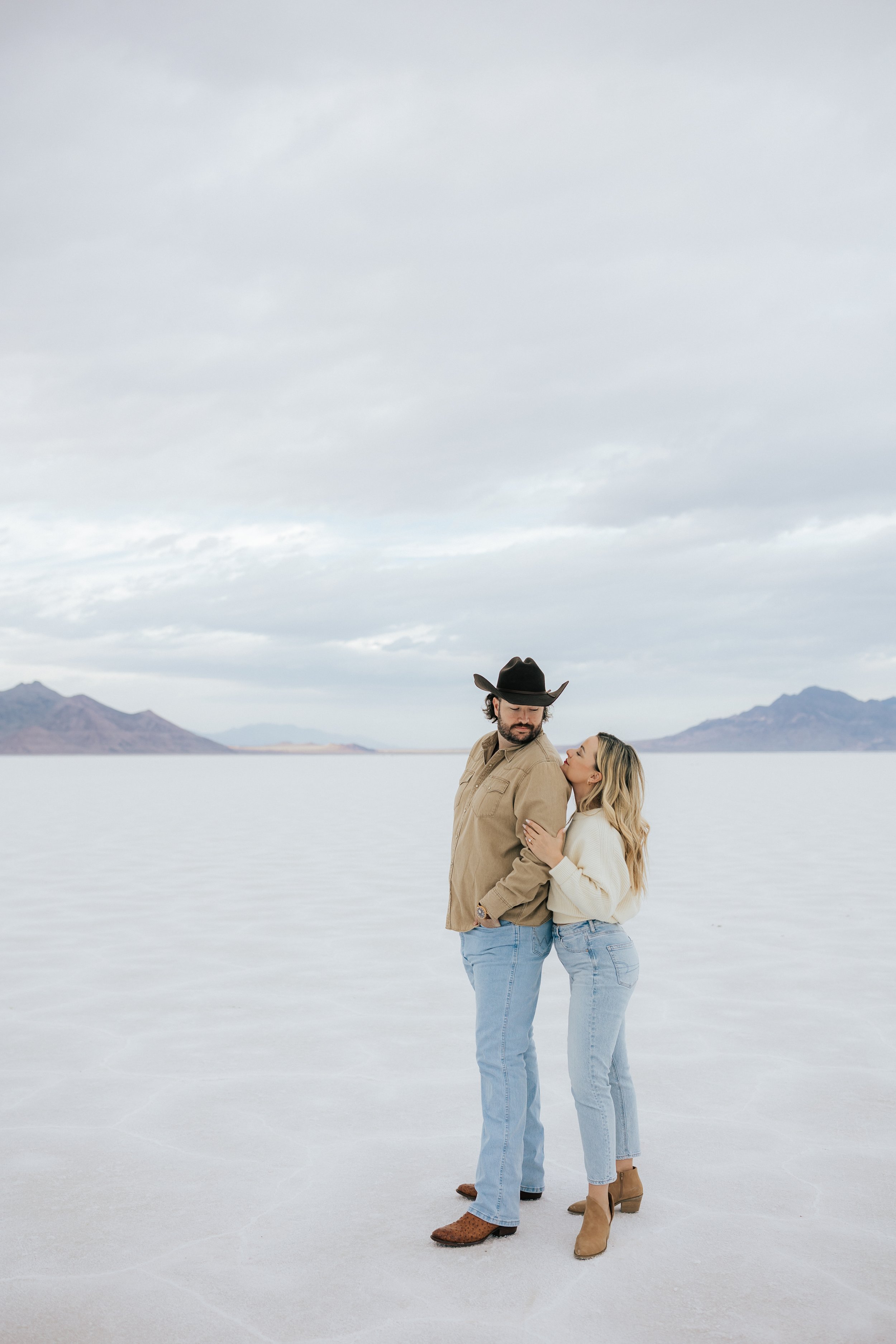  An engaged couple looks at each other as they pose for a photo at the Bonneville Salt Flats near Wendover, Nevada and Salt Lake City, Utah. The Salt Flats are white and the sky is overcast. The mountains show behind. Engagement session at the Utah S