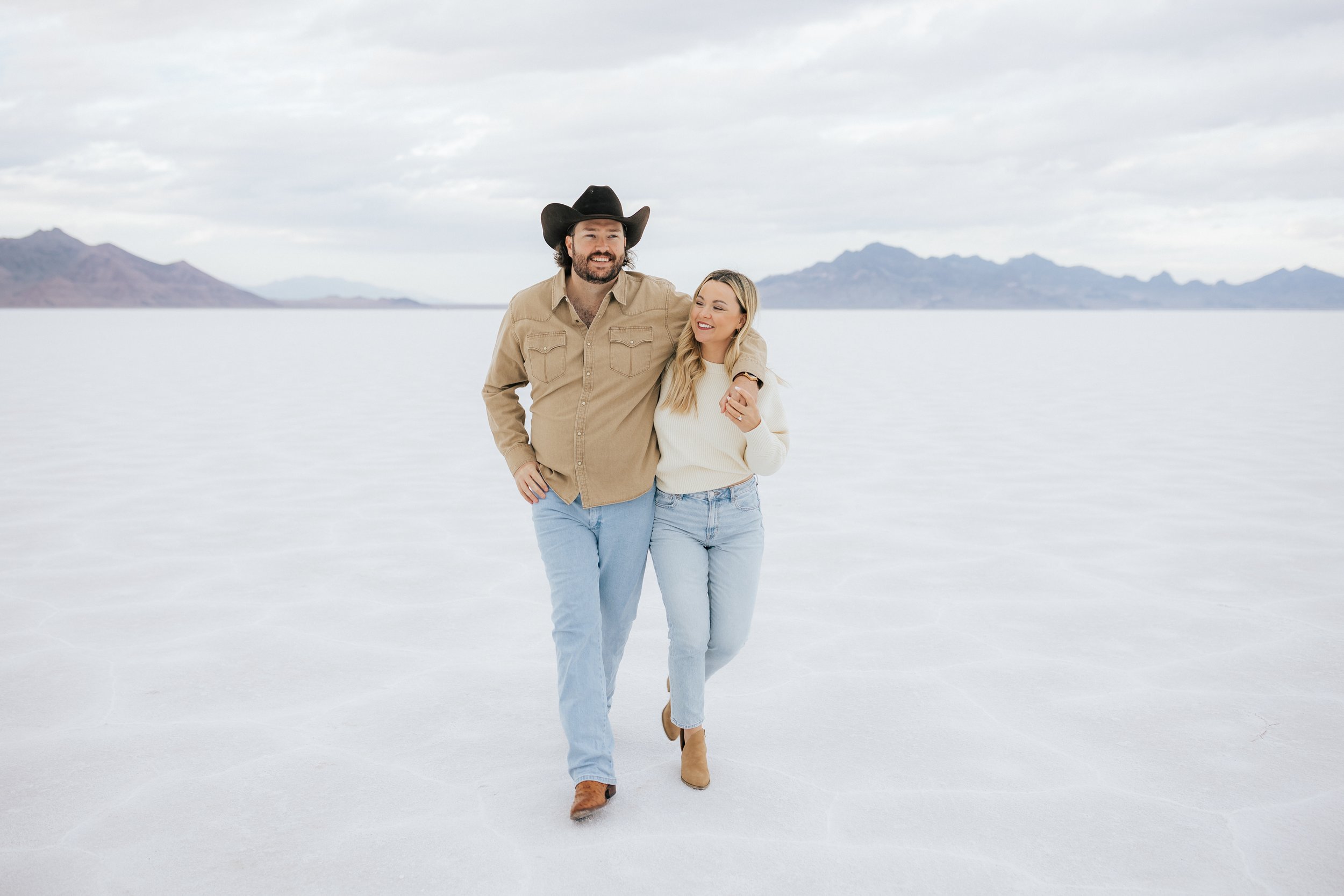 Photo of an engaged couple as they kiss and hug at the Bonneville Salt Flats near Wendover, Nevada and Salt Lake City, Utah. Cowboy and his girl walk together with his arm around her shoulder. The Salt Flats are white and the sky is overcast. The mo
