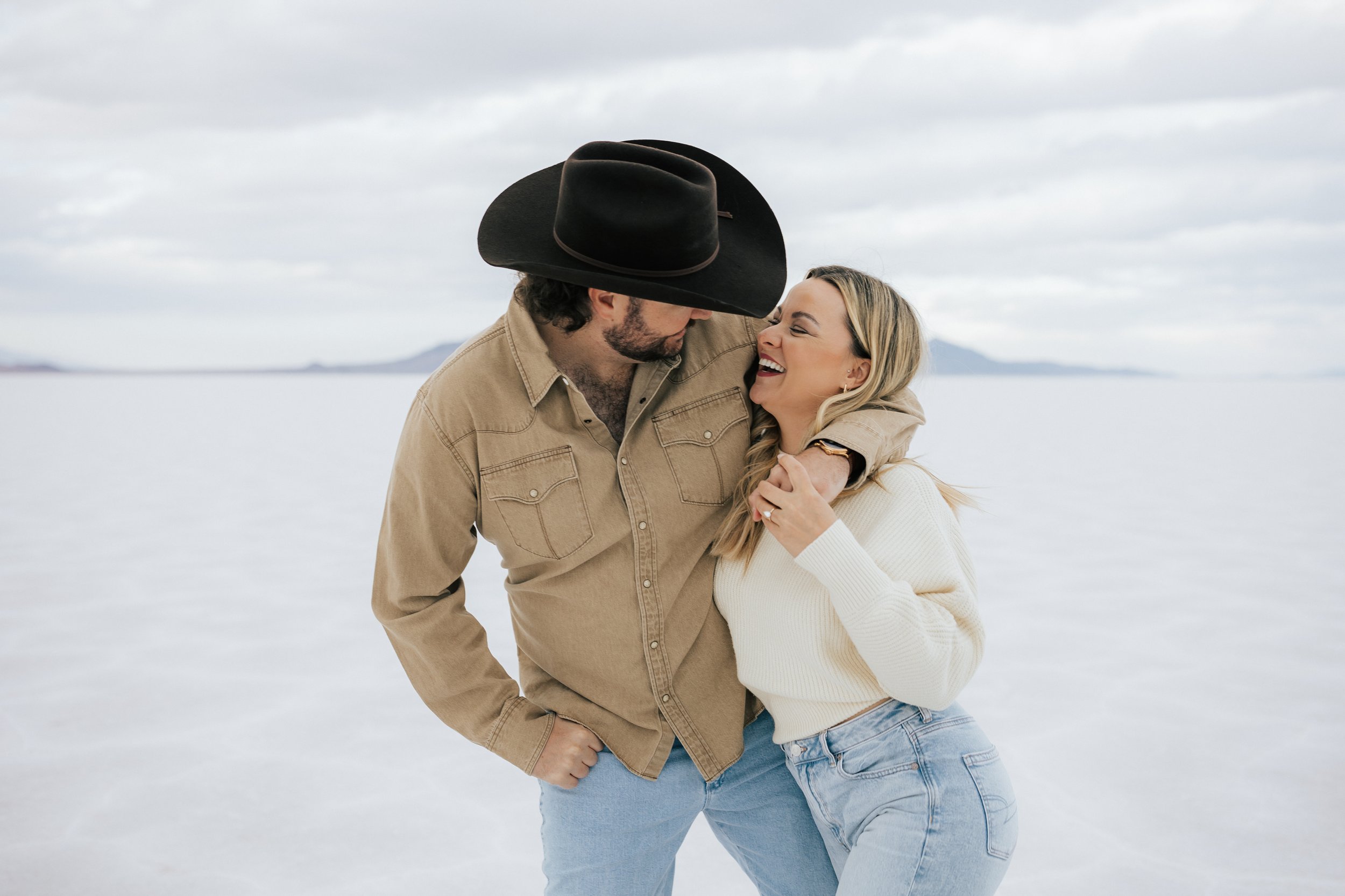  Photo of an engaged couple as they kiss and hug at the Bonneville Salt Flats near Wendover, Nevada and Salt Lake City, Utah. Cowboy and his girl walk together with his arm around her shoulder. The Salt Flats are white and the sky is overcast. The mo