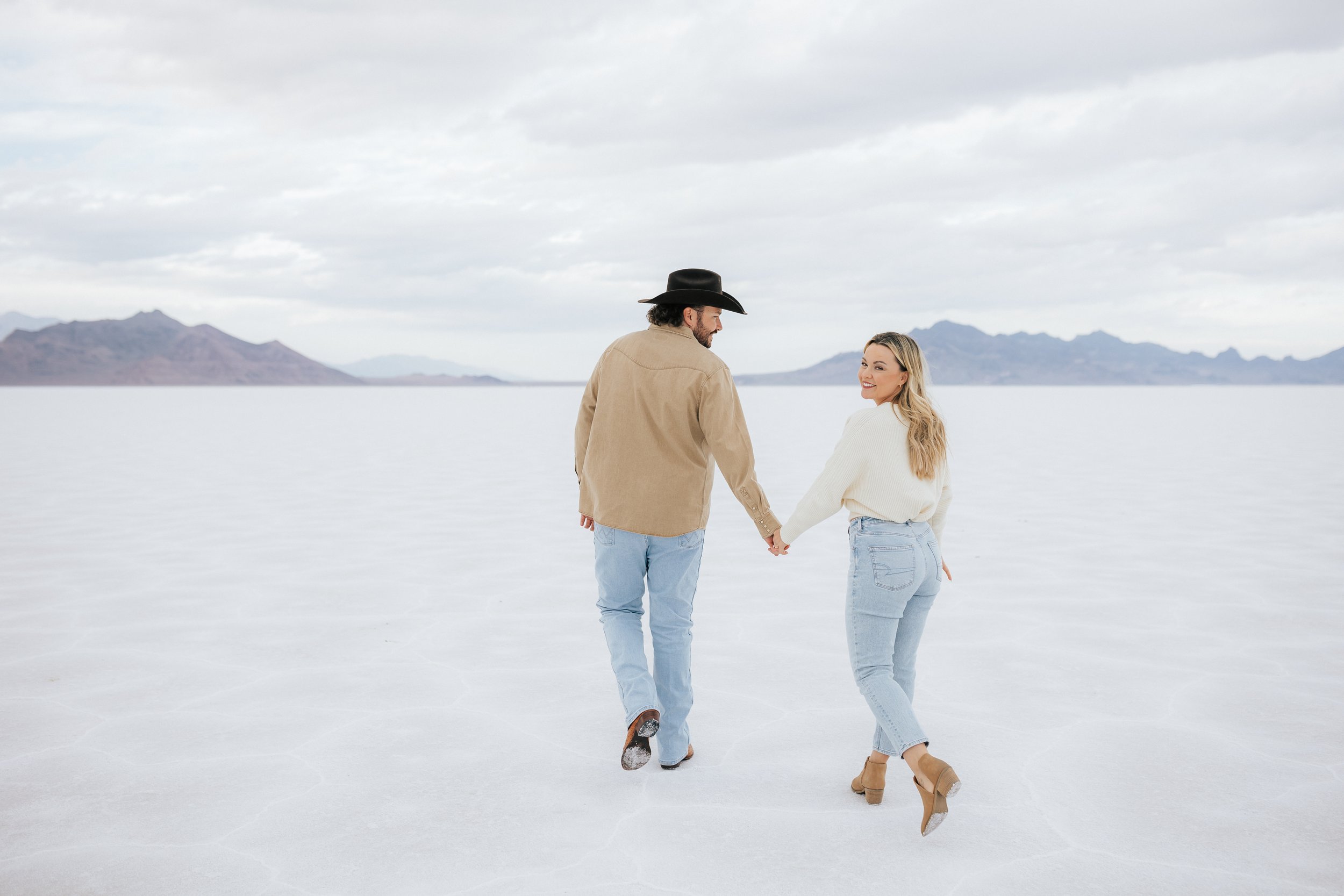   Photo of an engaged couple as they kiss and hug at the Bonneville Salt Flats near Wendover, Nevada and Salt Lake City, Utah. Cowboy and his girl walk together holding hands. The Salt Flats are white and the sky is overcast. The mountains show behin