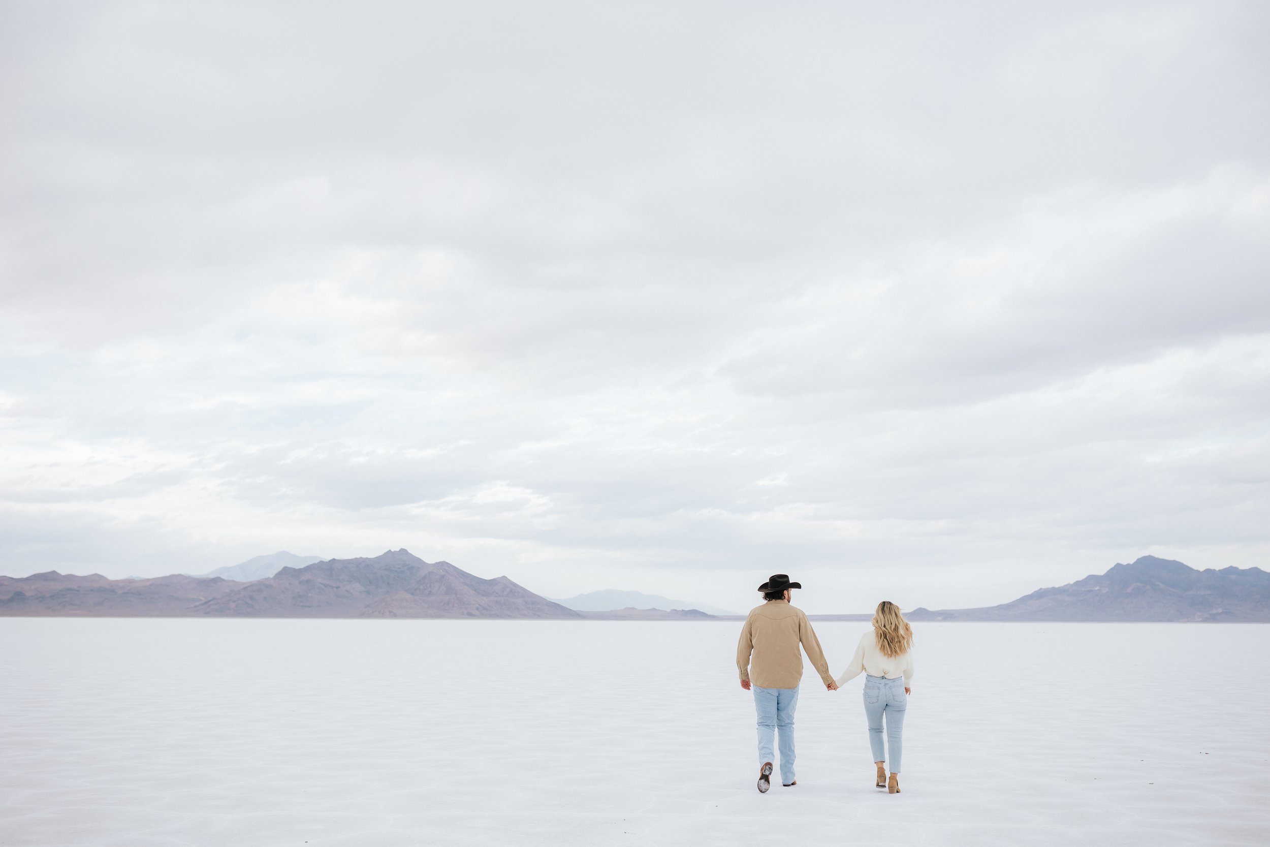  Photo of an engaged couple as they kiss and hug at the Bonneville Salt Flats near Wendover, Nevada and Salt Lake City, Utah. Cowboy and his girl walk together holding hands. The Salt Flats are white and the sky is overcast. The mountains show behind