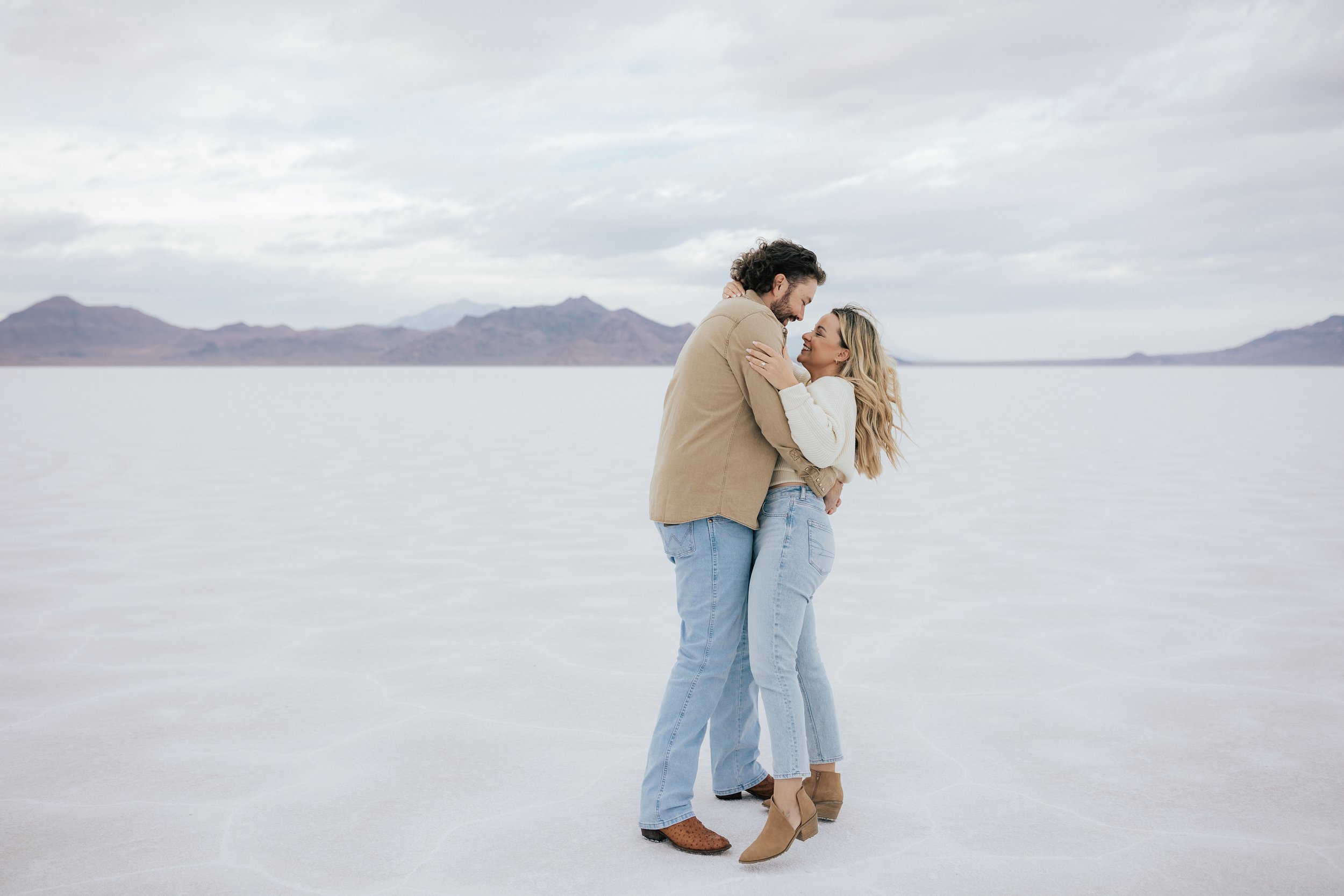  Engaged couple dance and kiss and laugh together at the Bonneville Salt Flats near Wendover, Nevada and Salt Lake City, Utah. Cowboy and his girl walk together with his arm around her shoulder. The Salt Flats are white and the sky is overcast. The m