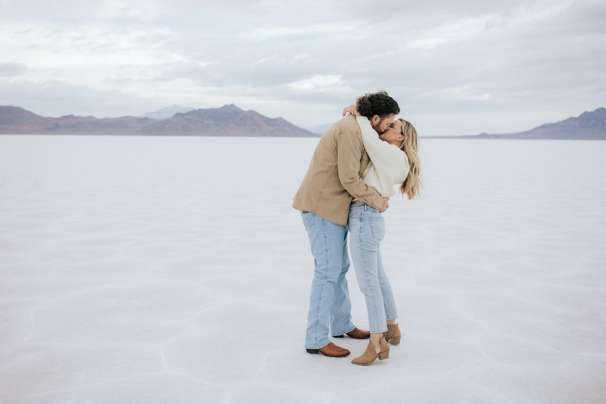  Photo of an engaged couple dancing as they kiss and hug at the Bonneville Salt Flats near Wendover, Nevada and Salt Lake City, Utah. Cowboy and his girl walk together with his arm around her shoulder. The Salt Flats are white and the sky is overcast