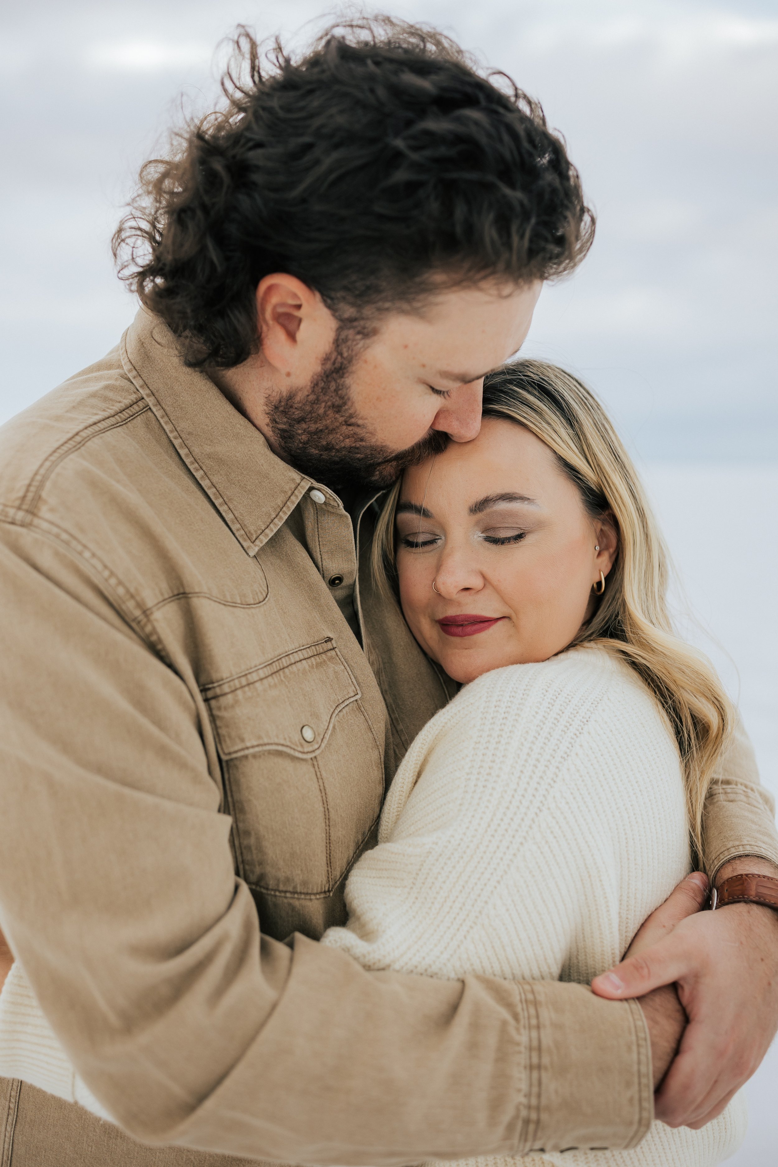  Engaged couple snuggles in a tight hug while he kisses her forehead at the Bonneville Salt Flats near Wendover, Nevada and Salt Lake City, Utah. The Salt Flats are white and the sky is overcast. The mountains show behind. Engagement session at the U