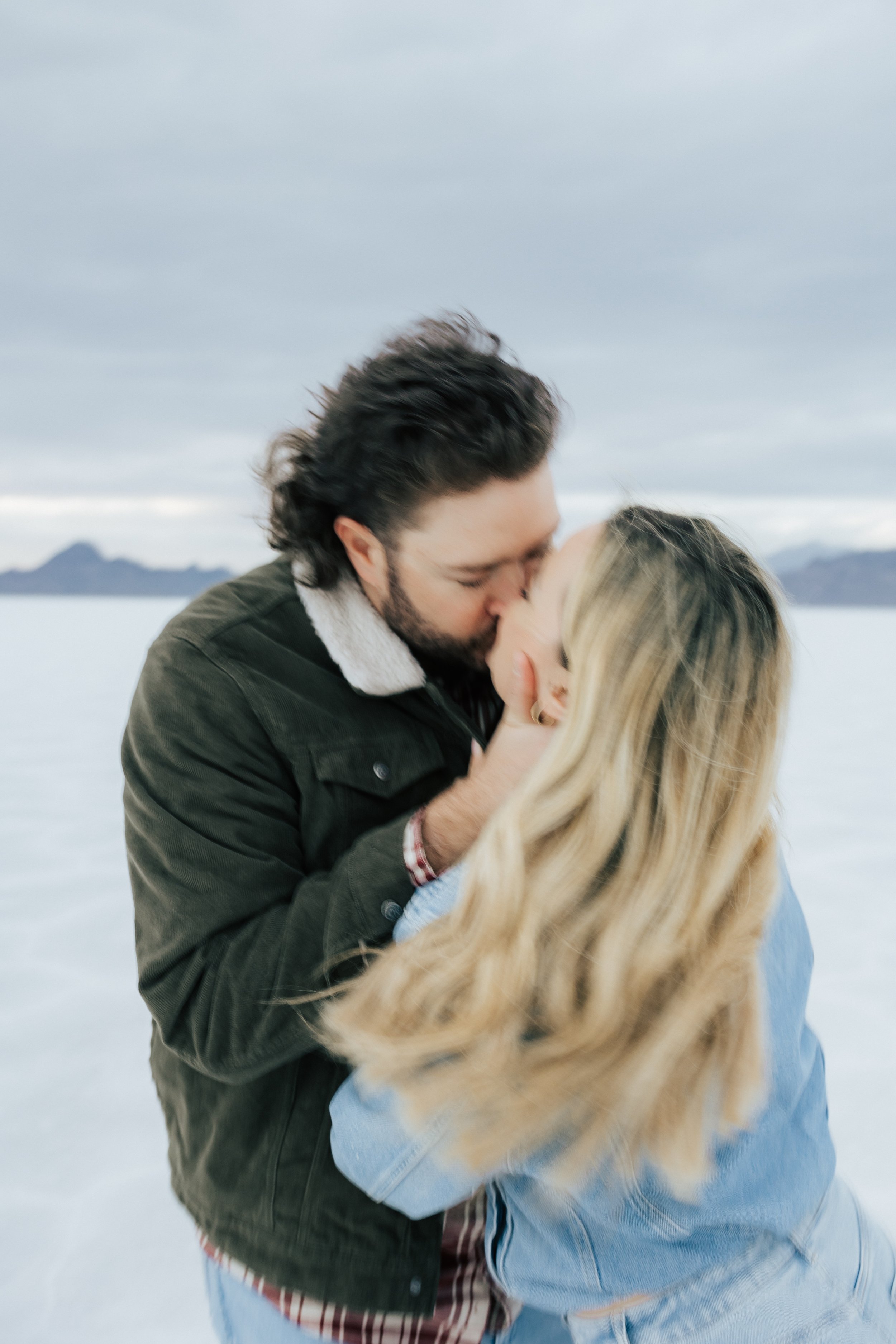  Blurry photo of an engaged couple as they kiss and hug at the Bonneville Salt Flats near Wendover, Nevada and Salt Lake City, Utah. The Salt Flats are white and the sky is overcast. The mountains show behind. Engagement session at the Utah Salt Flat