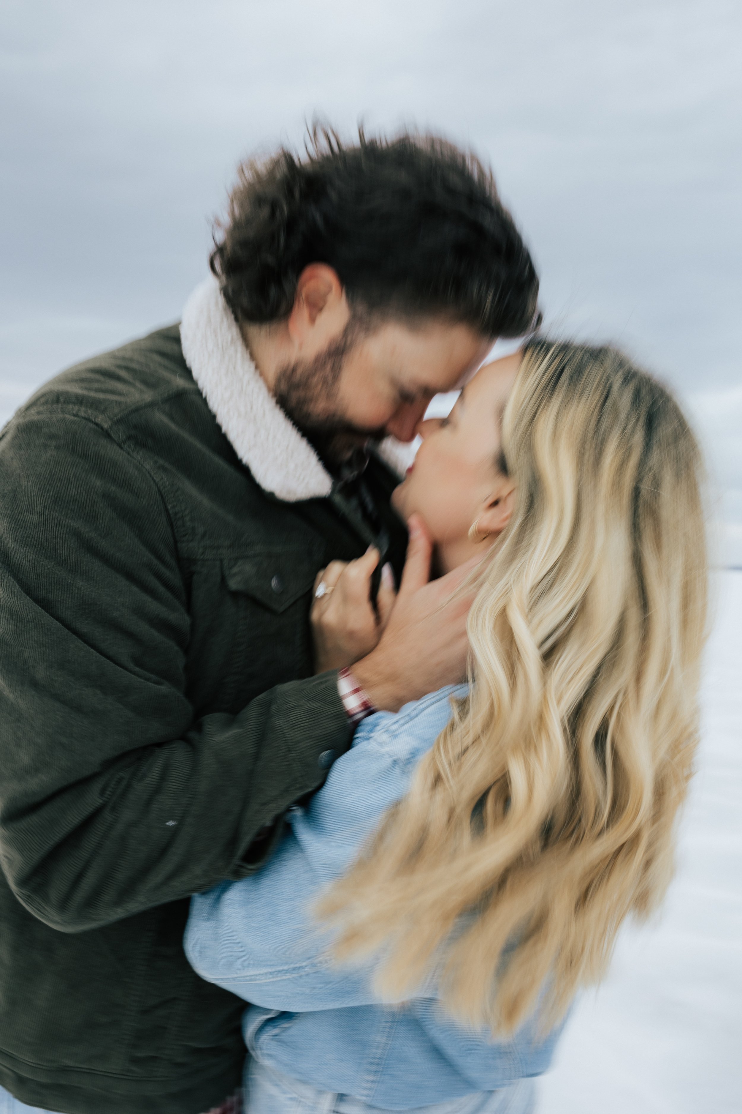  Blurry photo of an engaged couple as they hold each other and hug at the Bonneville Salt Flats near Wendover, Nevada and Salt Lake City, Utah. The Salt Flats are white and the sky is overcast. The mountains show behind. Engagement session at the Uta