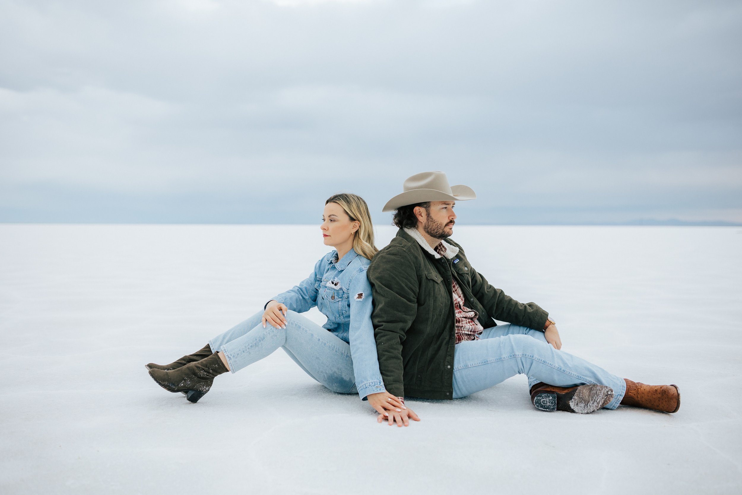 Engaged couple sits back to back on the salt at the Bonneville Salt Flats near Wendover, Nevada and Salt Lake City, Utah. The Salt Flats are white and the sky is overcast. The mountains show behind. Engagement session at the Utah Salt Flats. Salt Fl