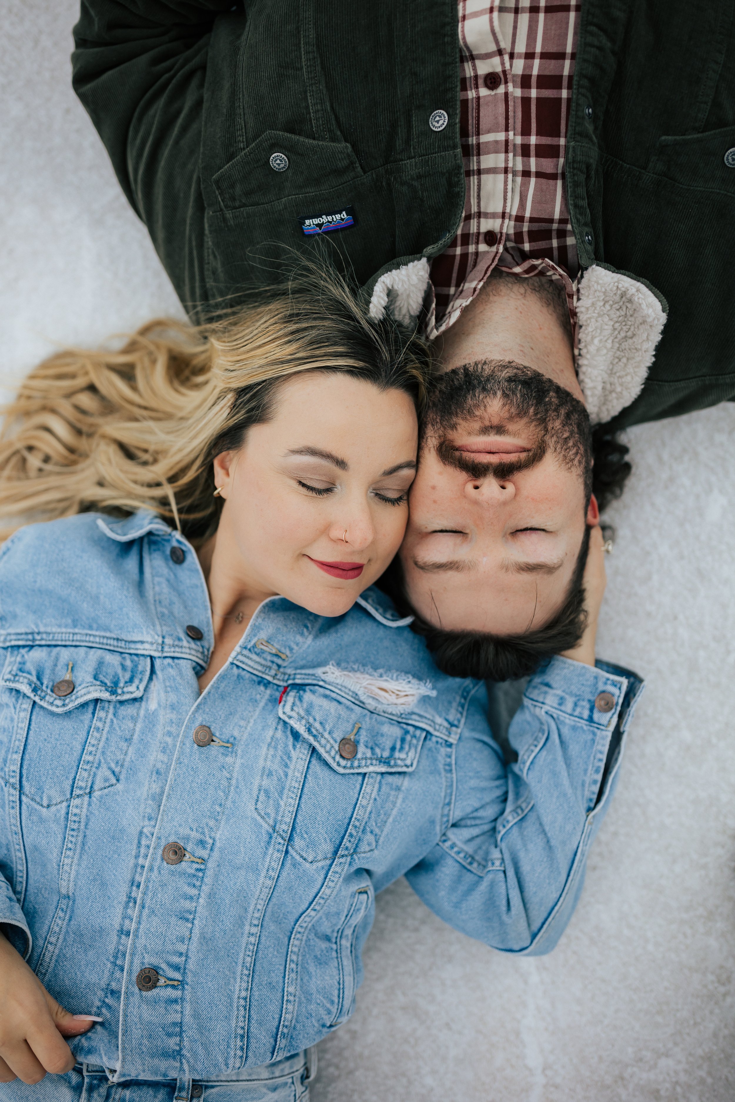  An engaged couple lays down together in opposite directions with their heads touching on the Bonneville Salt Flats near Wendover, Nevada and Salt Lake City, Utah. The girl is wearing a blue denim jacket and the man is wearing a plaid shirt with a co