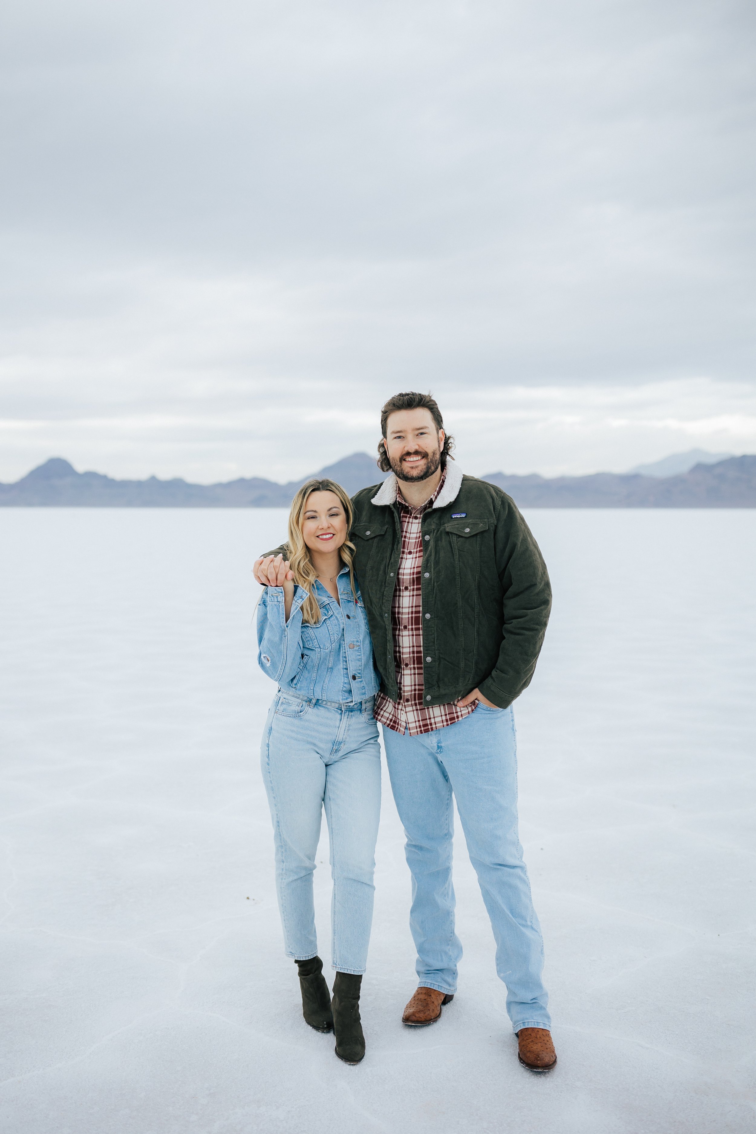  An engaged couple smiles as they pose for a photo at the Bonneville Salt Flats near Wendover, Nevada and Salt Lake City, Utah. They are both wearing jackets and jackets. Engagement session at the Utah Salt Flats. #engagements #utahphotographer 