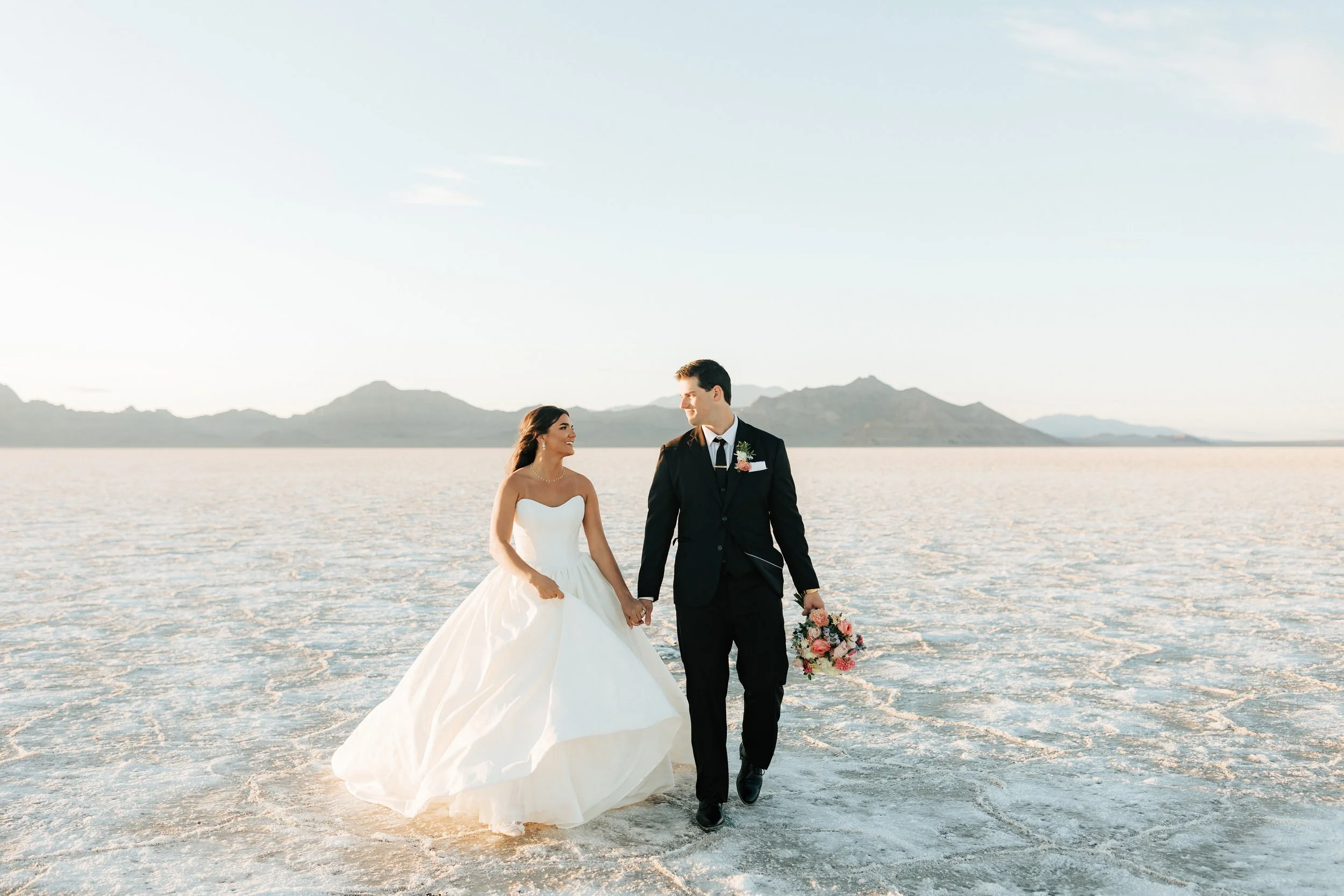  Bride and groom walk together at the Salt Flats at sunset in their wedding dress and suit. 